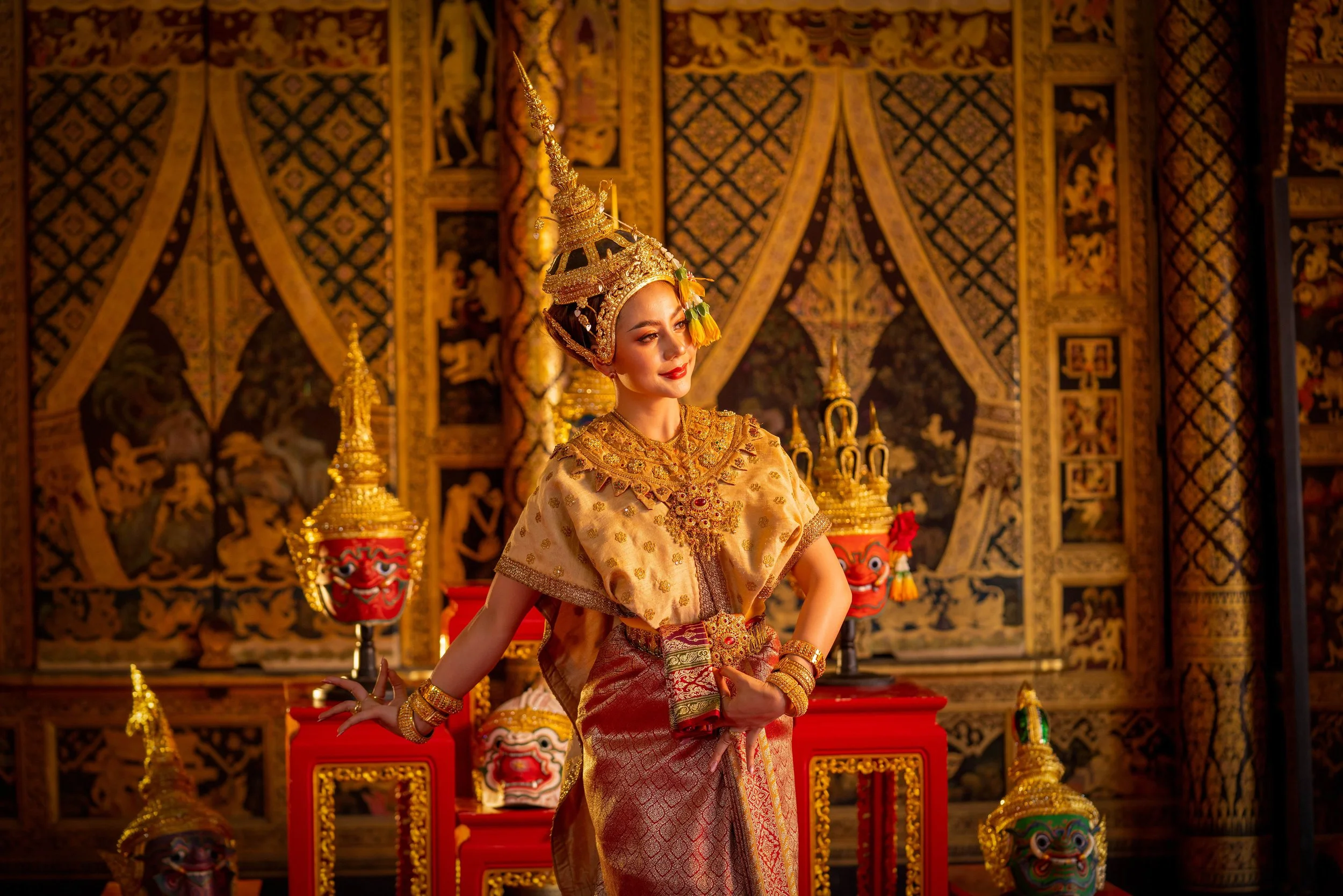 A woman dressed in traditional Thai attire with elaborate gold accessories and headpiece, standing in front of ornate, decorative backgrounds with ceremonial masks and sculptures.
