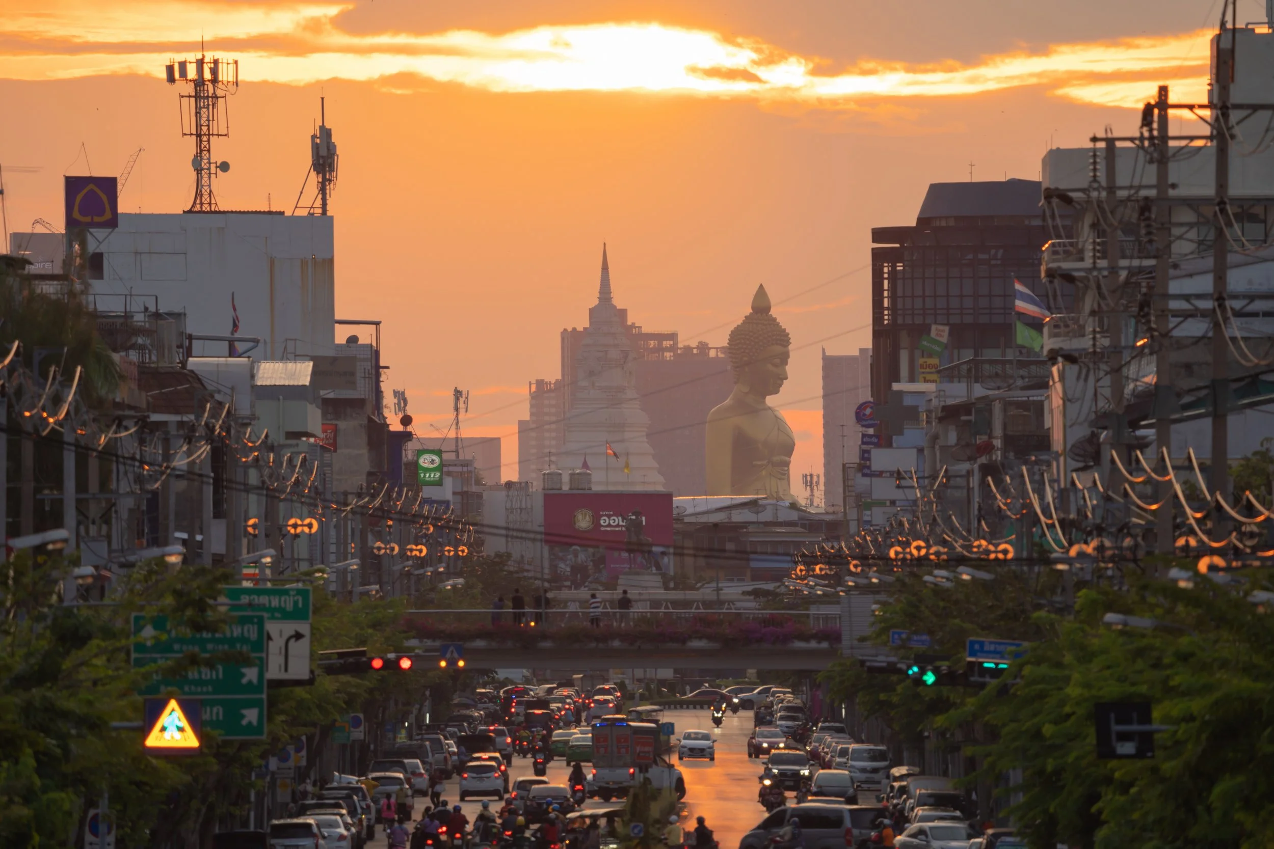 City street at sunset with cars, motorcycles, and traffic lights, large Buddha statues and tall buildings in the background, and a partly cloudy sky.