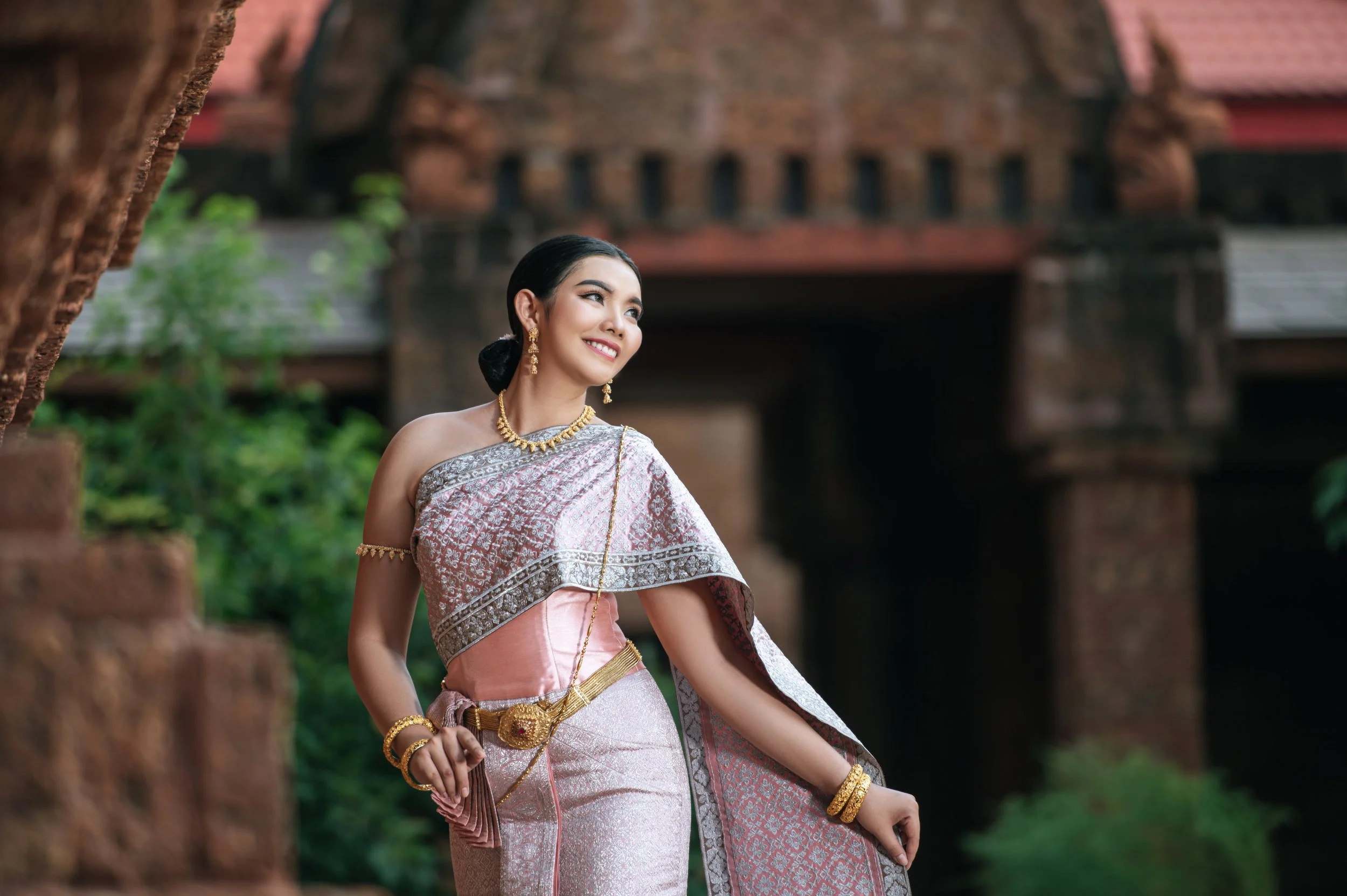 A woman in traditional Thai attire standing outdoors with ancient brick architecture and greenery in the background.