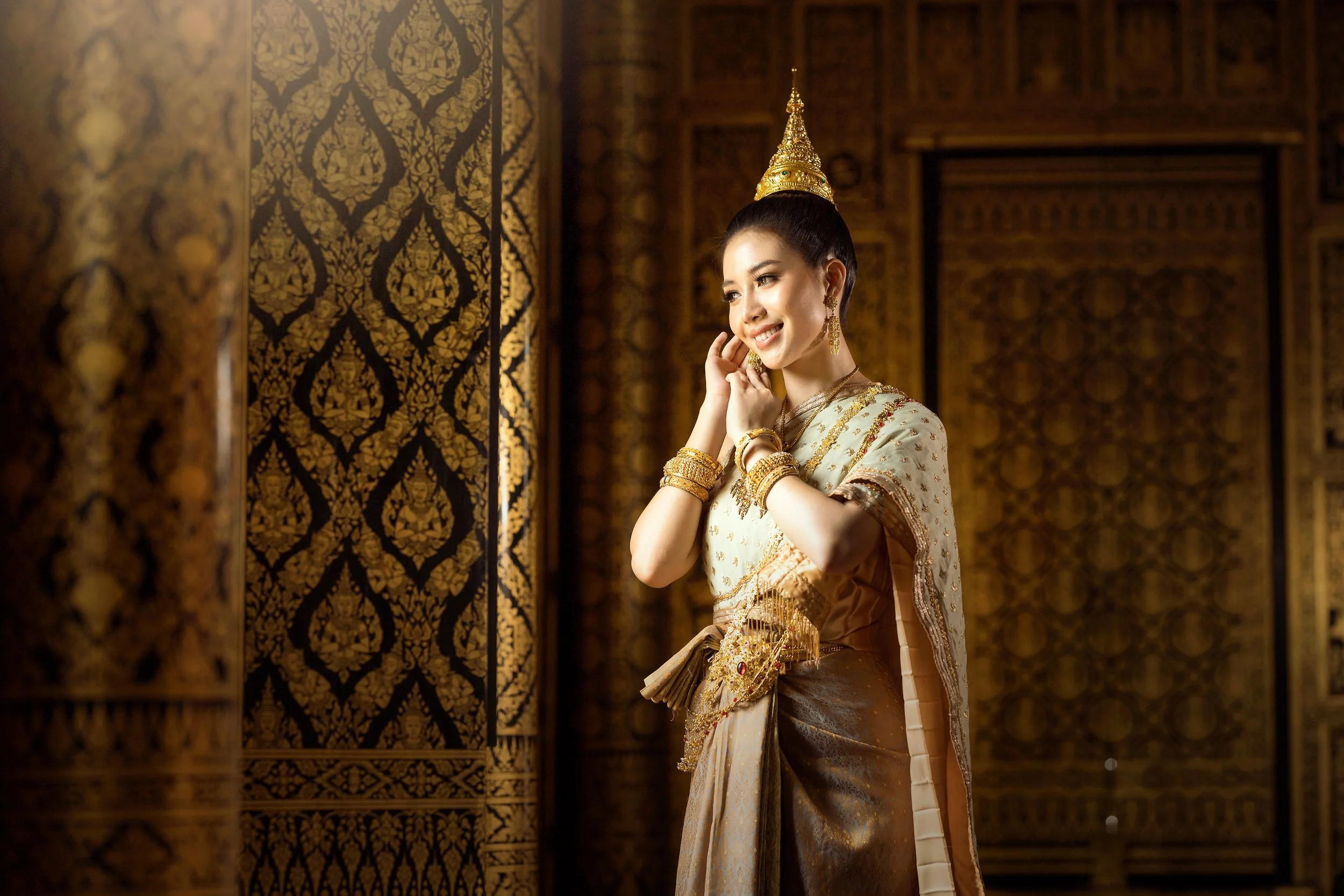 A woman in traditional Thai clothing and gold jewelry smiling and touching her face, standing in front of ornately decorated wooden doors.