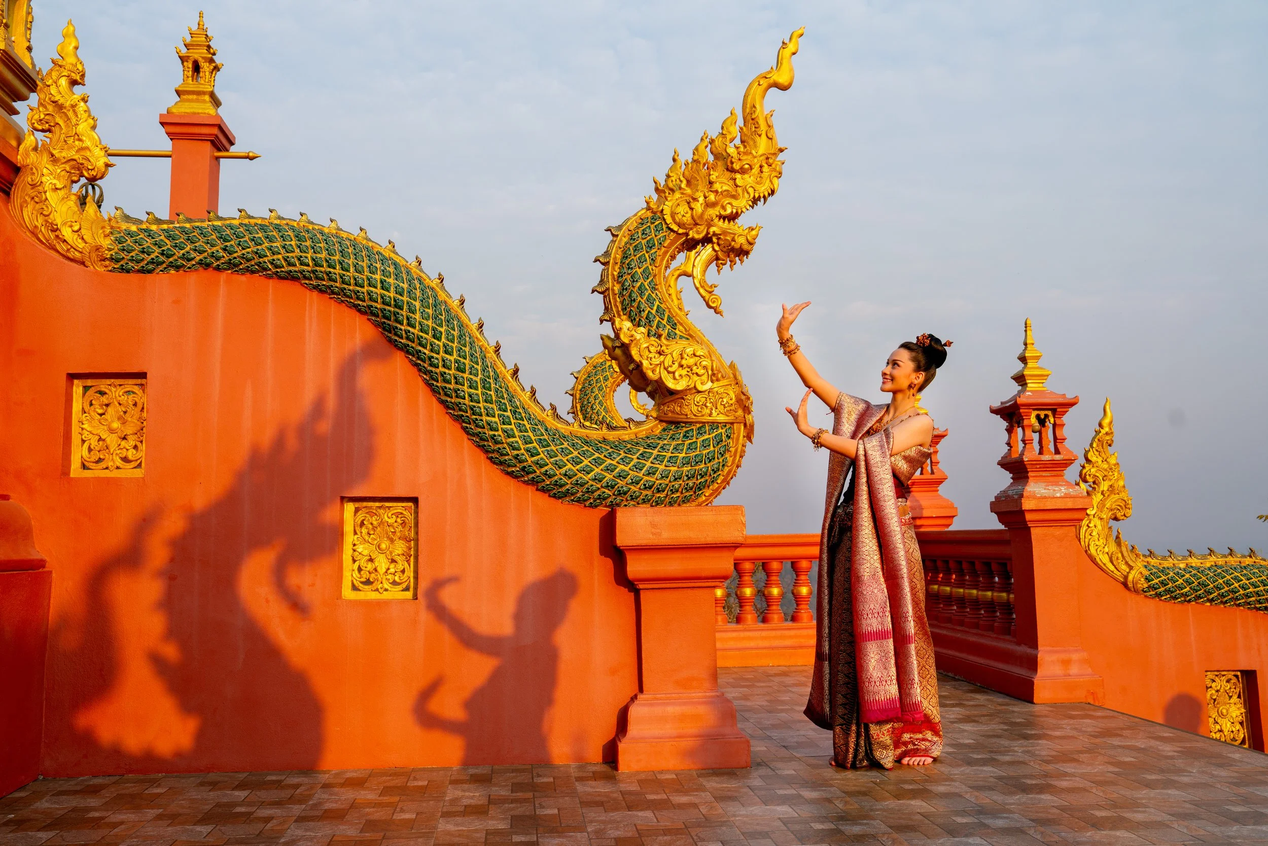 A woman in traditional Thai attire dancing on the orange and gold ornate terrace of a temple, with the shadow of her dance projected on the wall behind her.
