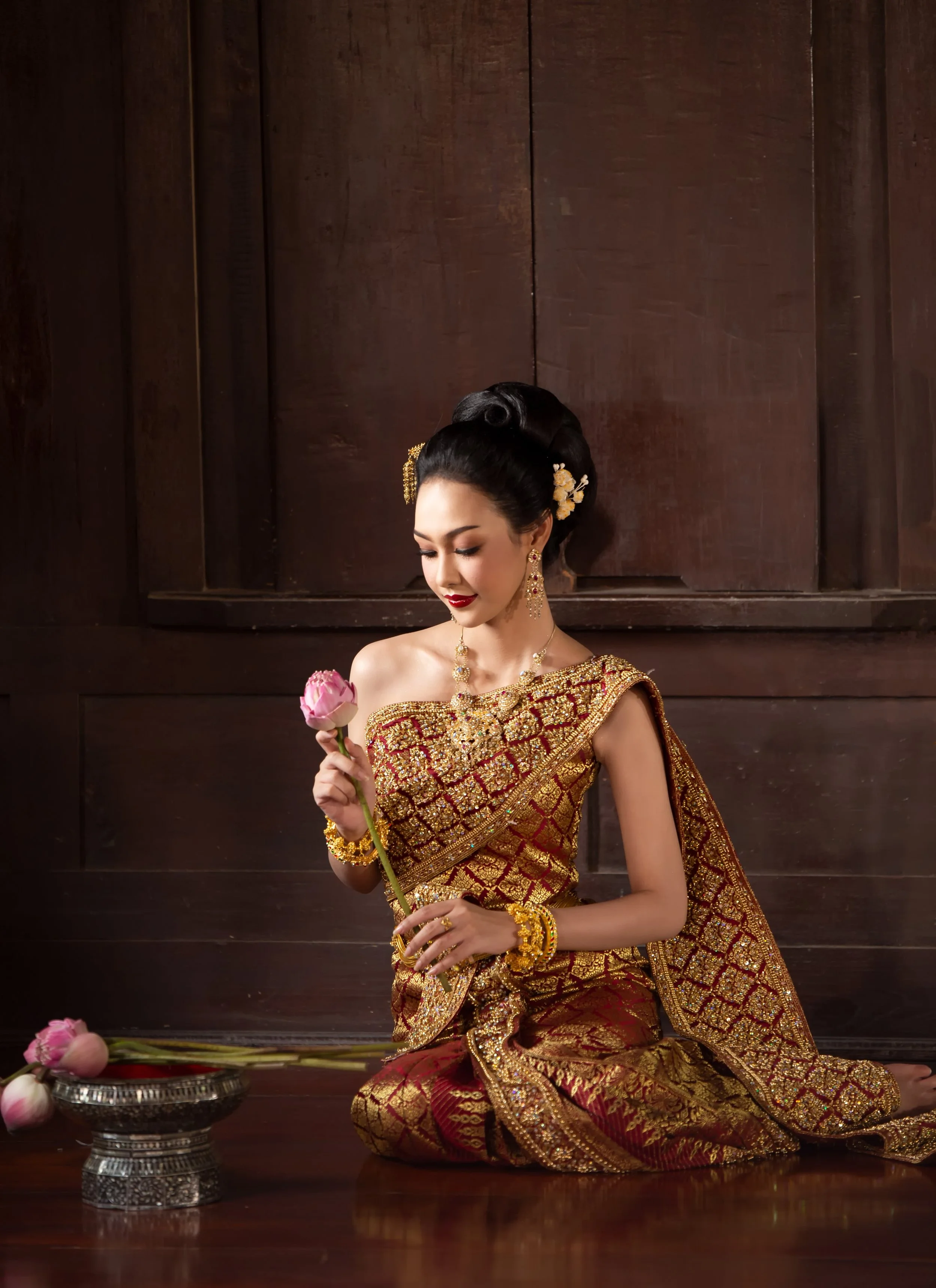 A woman dressed in traditional Thai attire sitting on the wooden floor, holding a pink lotus flower, with more lotus flowers placed on a silver tray beside her.