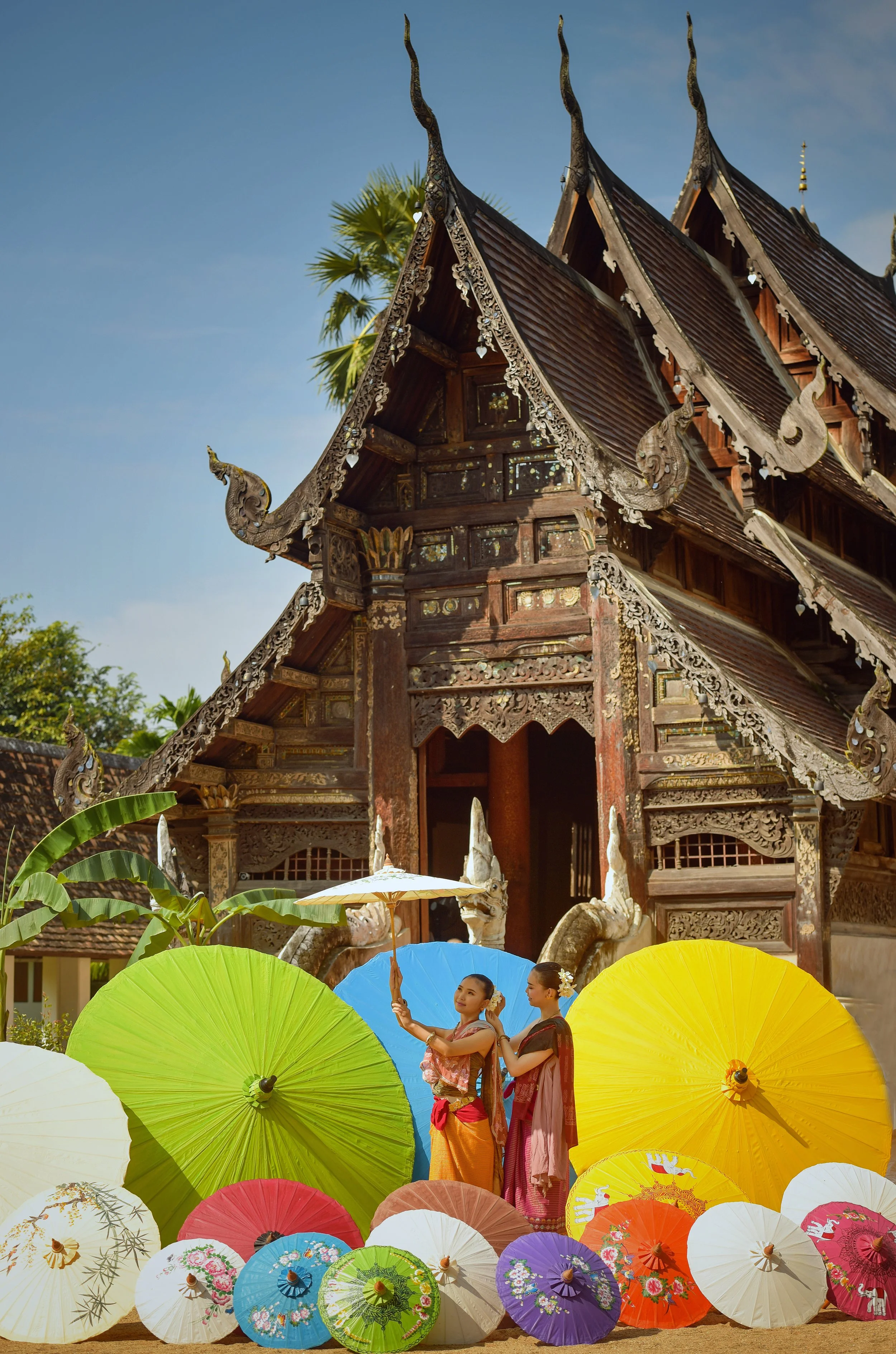 Two women in traditional Thai attire holding umbrellas in front of a temple with colorful paper umbrellas on the ground.