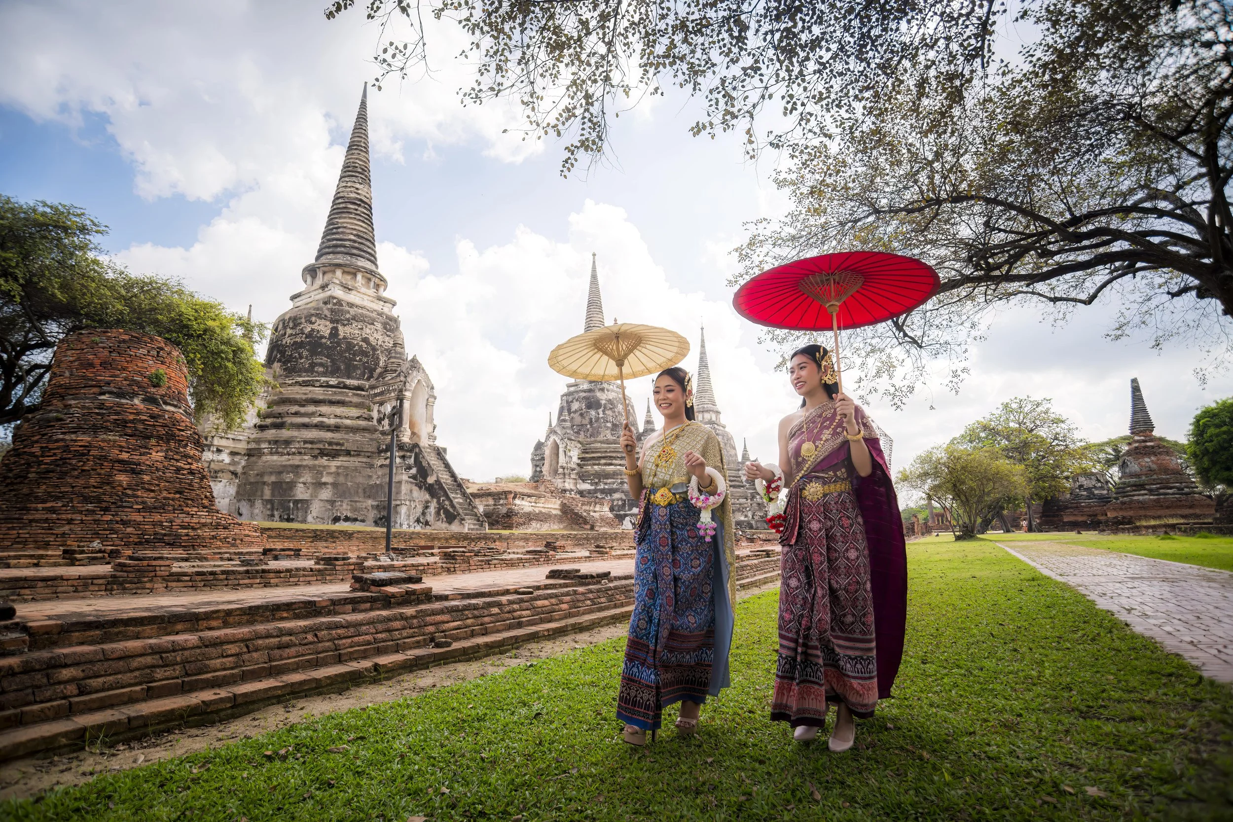 Two women in traditional Thai clothing walking on grass, holding umbrellas, near ancient ruins and pagodas in a park with trees and a cloudy sky.