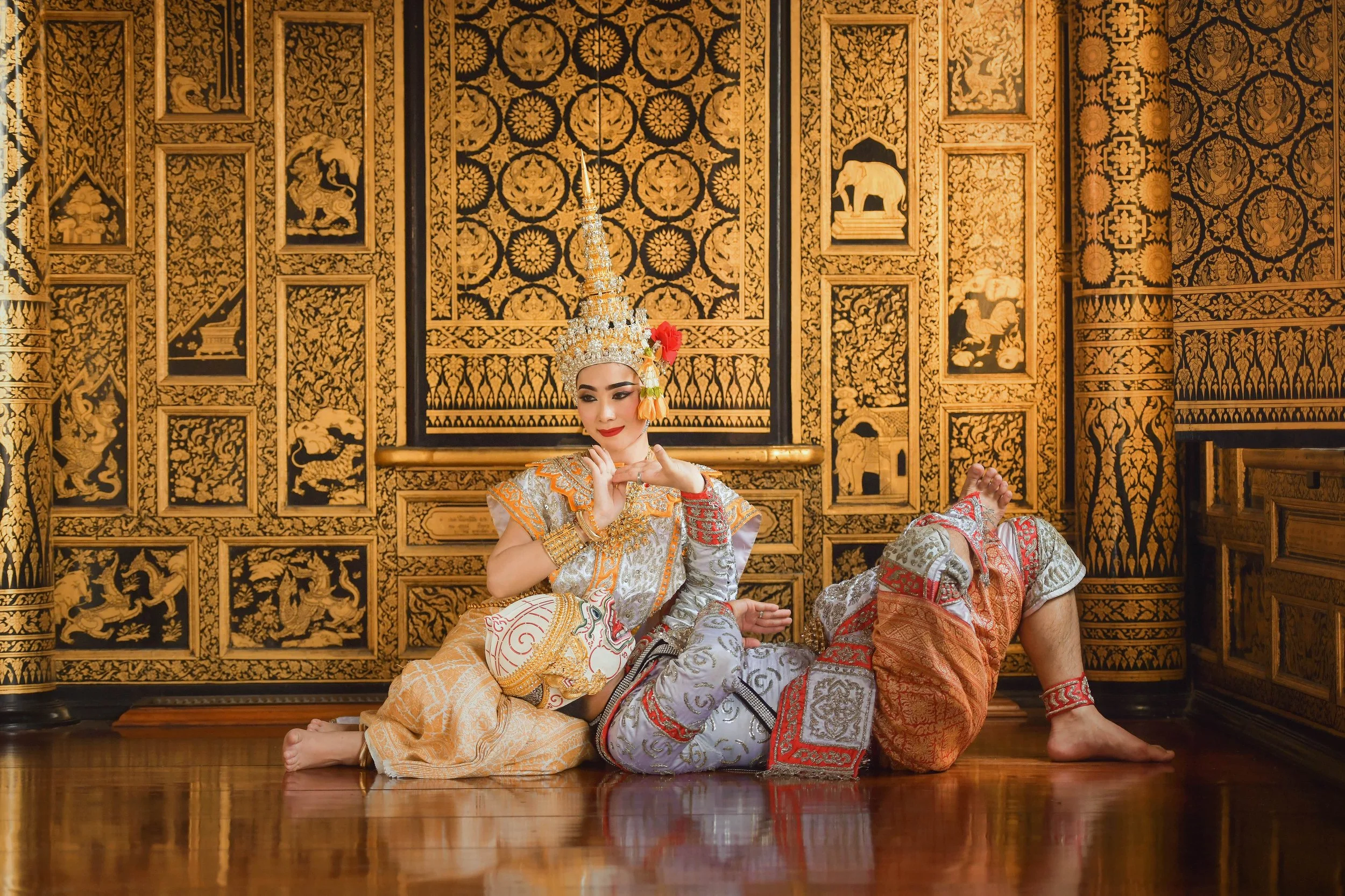 A woman dressed in traditional Thai costume, sitting on the floor in front of an ornate, gold and black carved wooden wall, decorated with intricate patterns and animal motifs, in a traditional Thai temple setting.