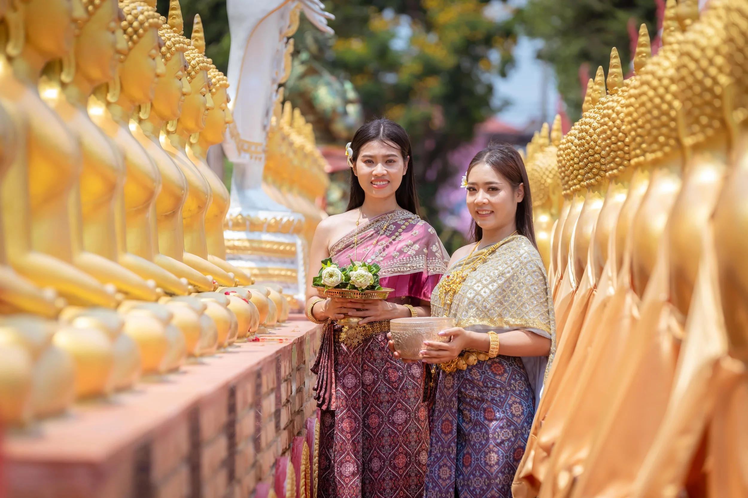 Two women dressed in traditional Thai clothing are standing outdoors at a temple or shrine, surrounded by golden Buddha statues. They are holding bowls with flowers and offerings, smiling at the camera.