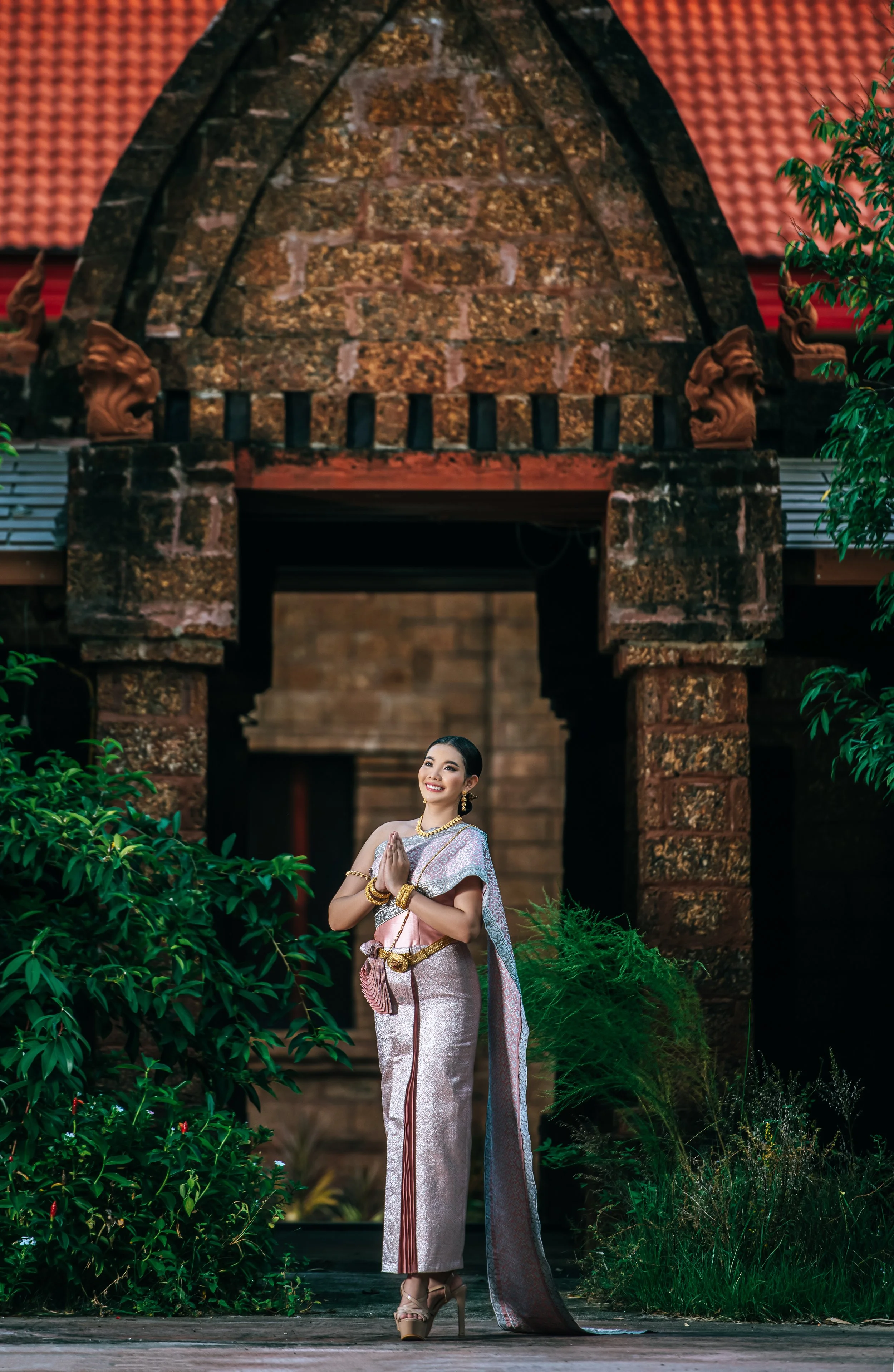 A woman dressed in traditional Thai attire, standing outdoors in front of an ornate archway with greenery around.