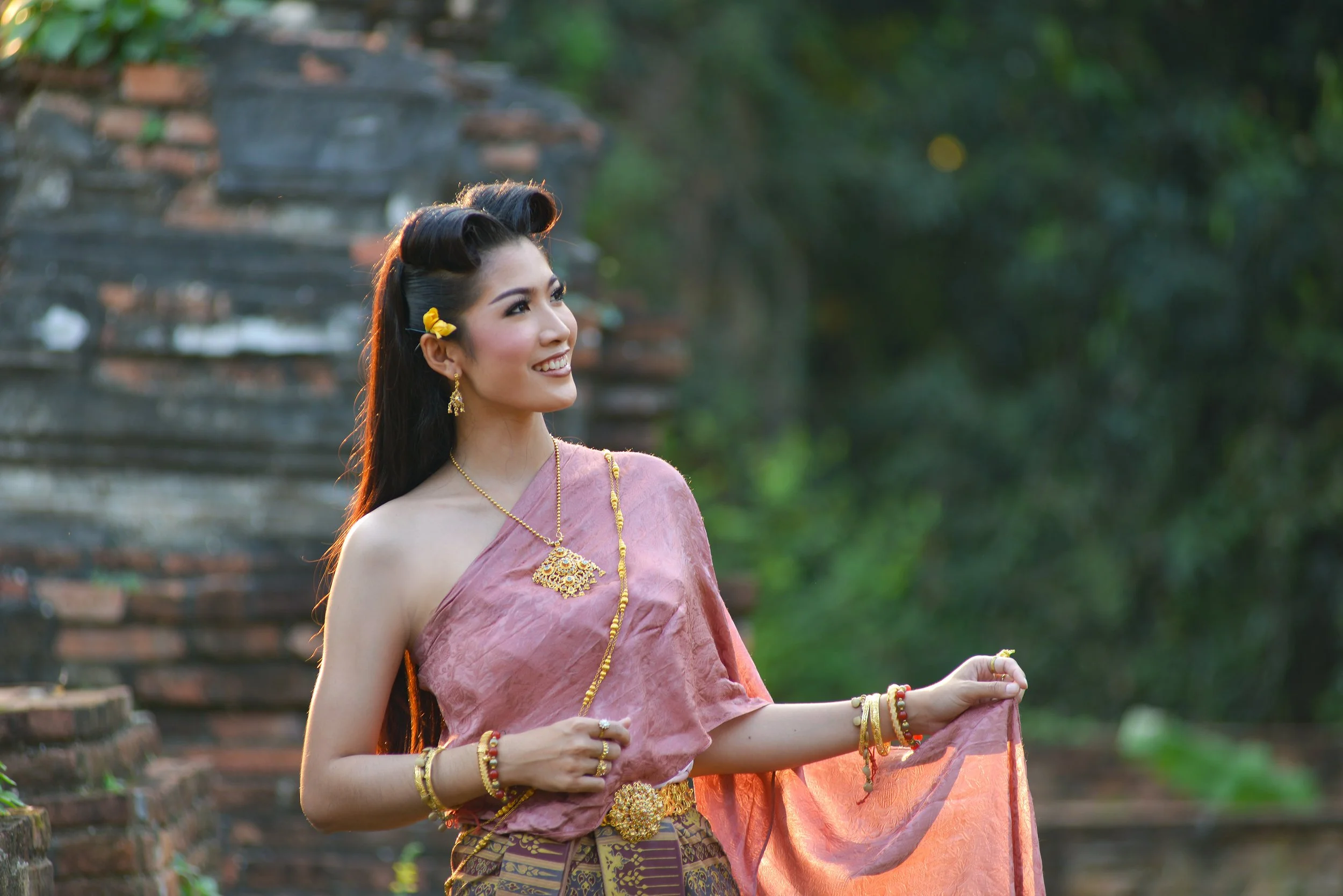 A young woman dressed in traditional Thai attire, wearing gold jewelry and smiling outdoors with trees and ancient brick ruins in the background.