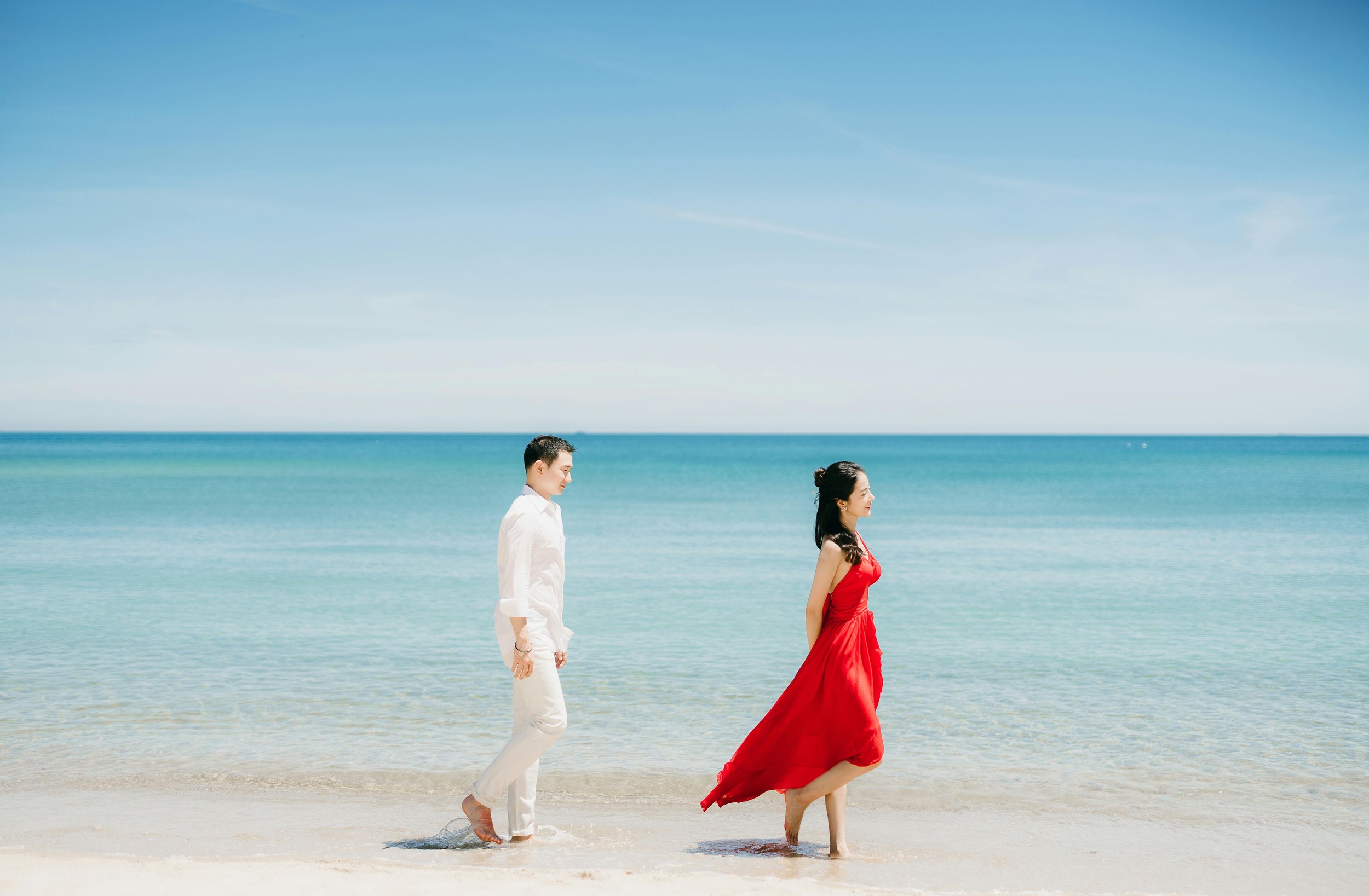 A man in a white shirt and white pants walking barefoot on the beach, followed by a woman in a red dress also barefoot, with the ocean and blue sky in the background.