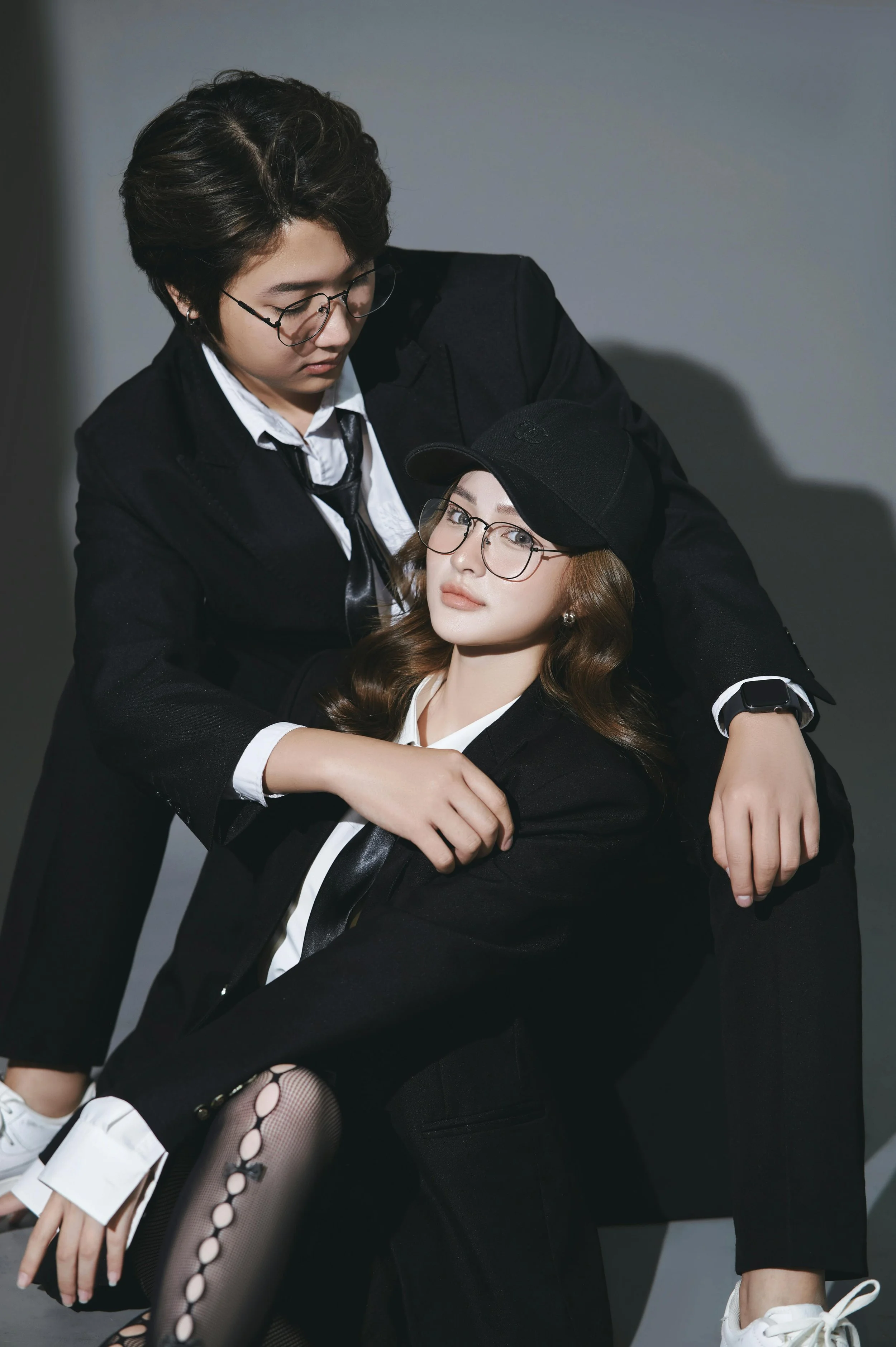 Two women wearing black suits and glasses posing together in a studio, with one standing and the other sitting, against a gray background.