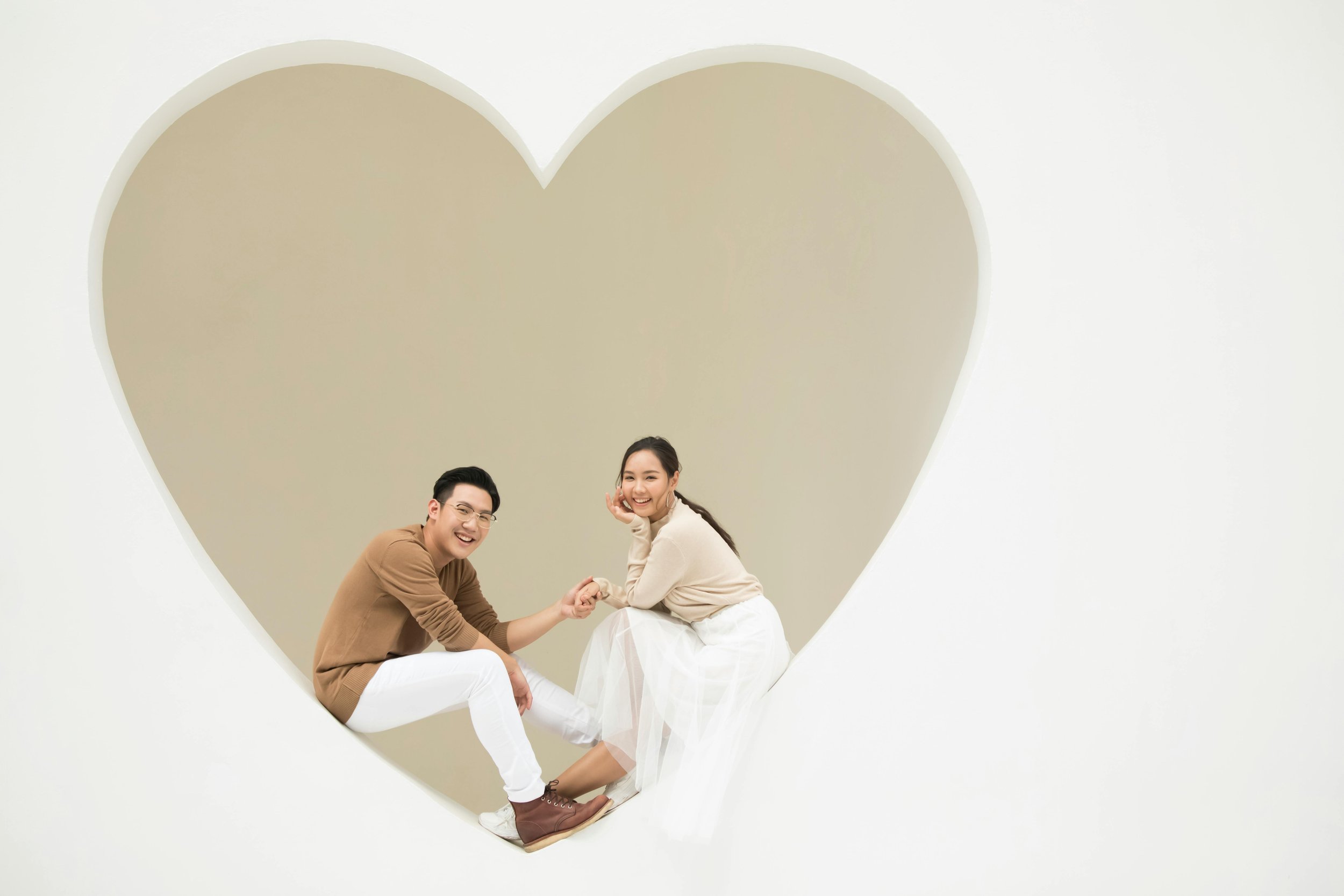 A smiling man and woman sitting inside a large heart-shaped cutout in a blank background.