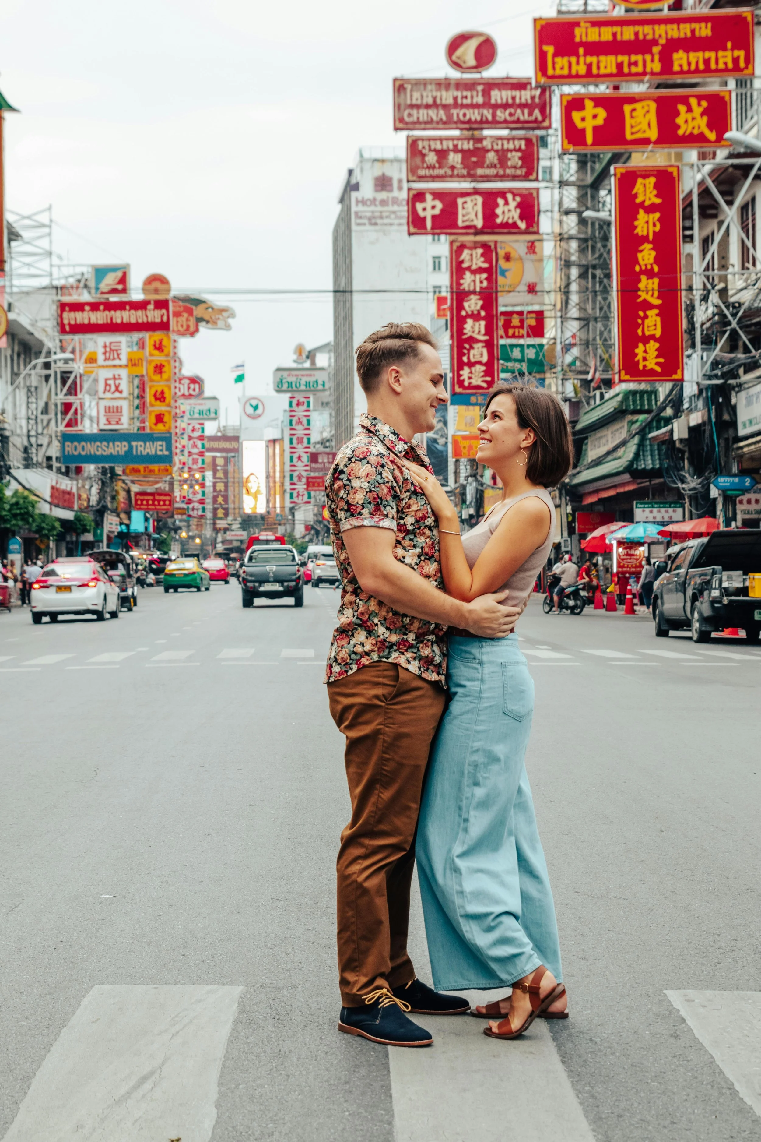 A couple standing and smiling affectionately in the middle of a busy city street with numerous colorful signs in Chinese and Thai languages.