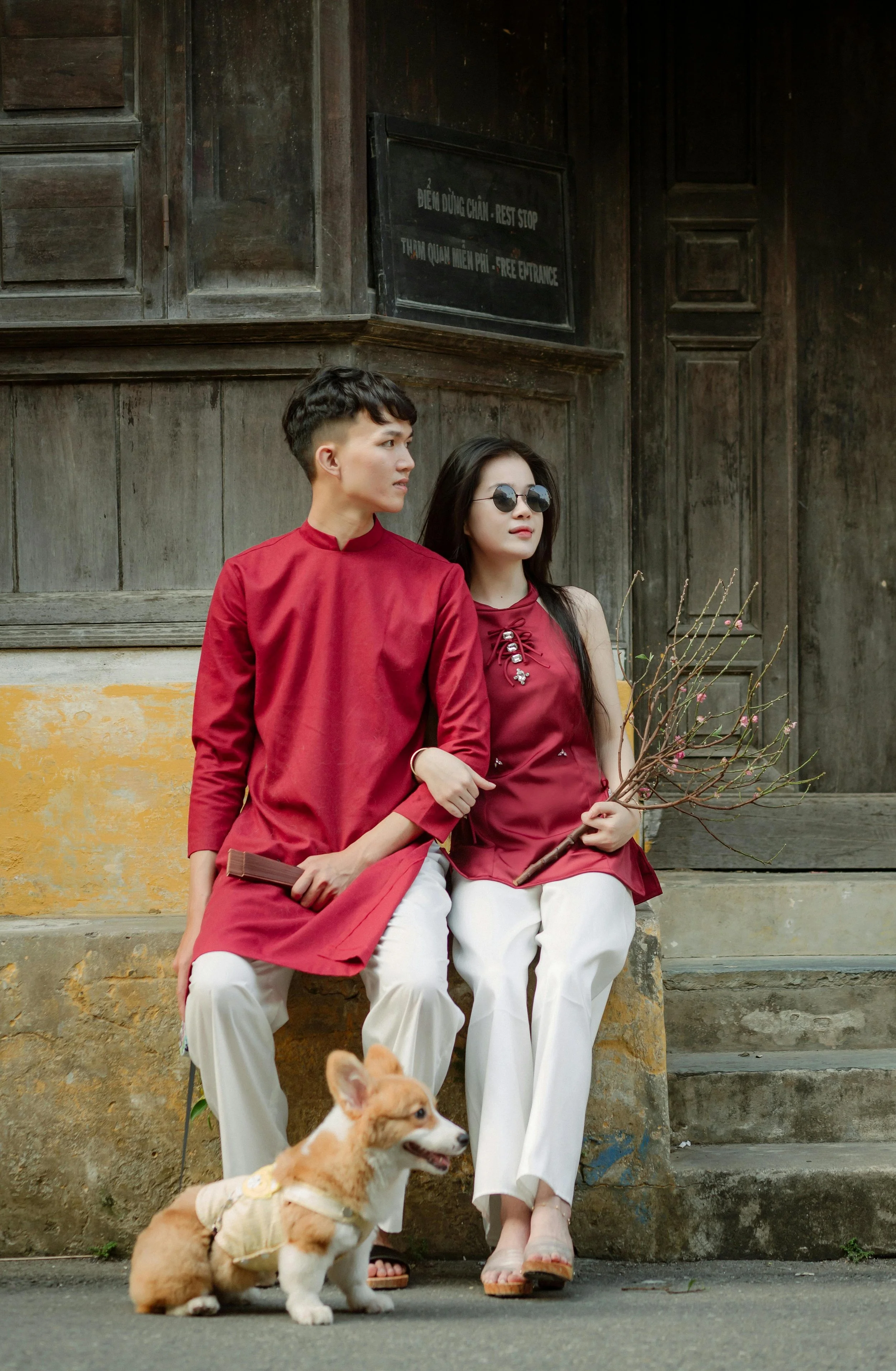 A young man and woman, both dressed in red tops and white pants, sit on a concrete step outside a wooden building. The woman is holding a branch with pink blossoms, and the man is holding a wooden object. A small fluffy dog with a beige fur coat, wea