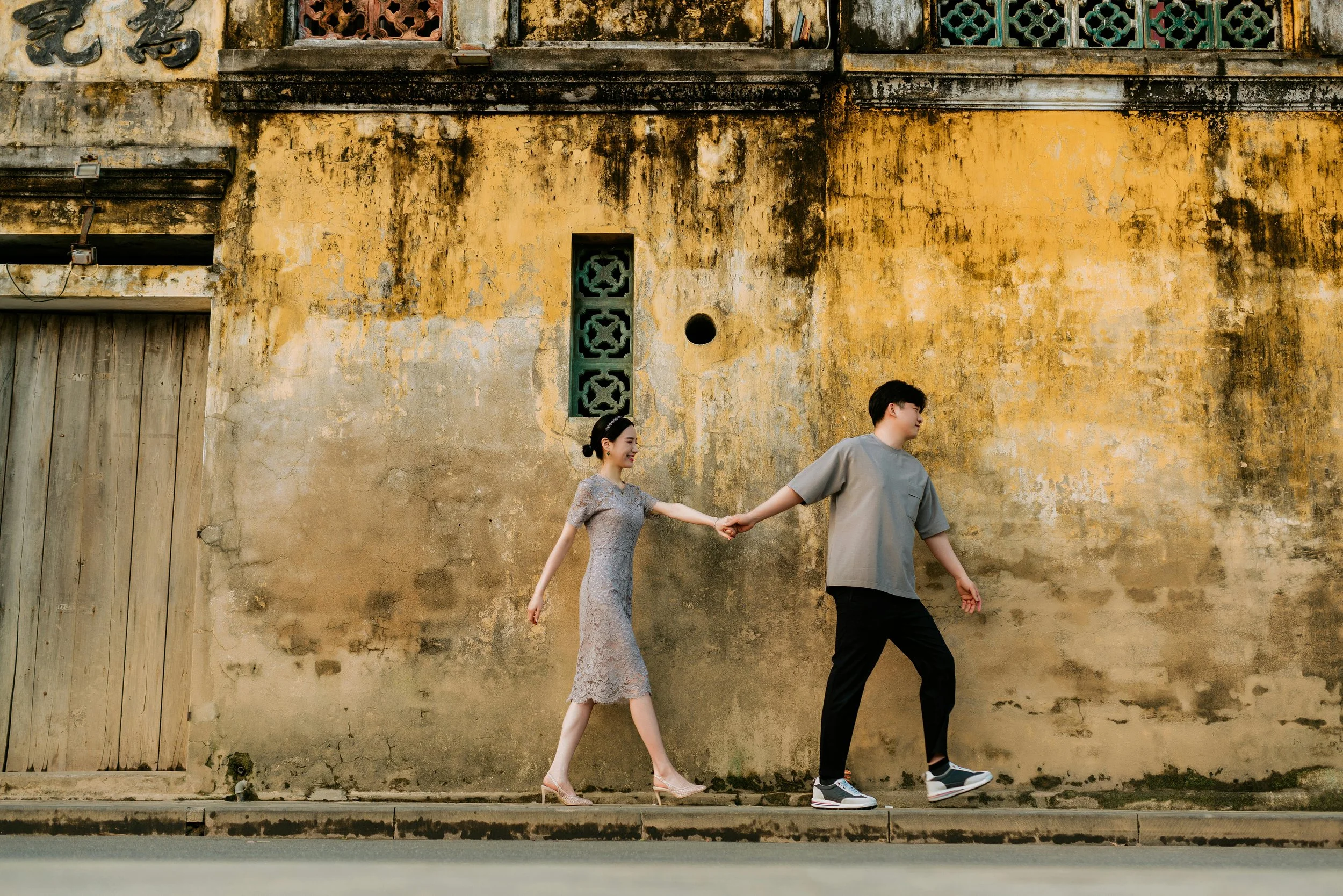 A woman in a gray lace dress holding hands with a man in a gray T-shirt and black pants walking along a weathered yellow wall.