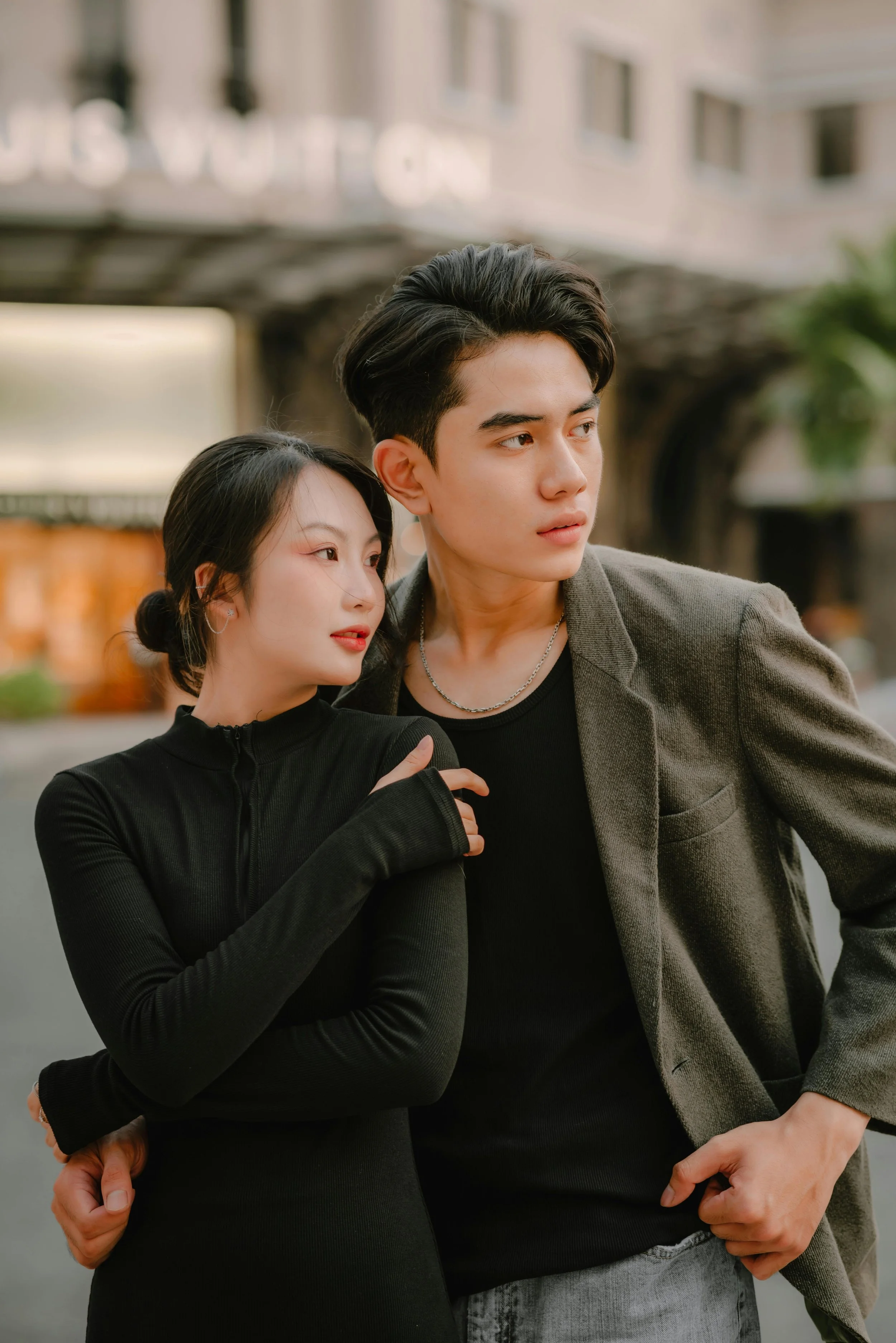 A young woman and a young man stand close together on a city street, with the woman resting her head on the man's shoulder and looking at him, both dressed in black, with buildings and blurred signs in the background.