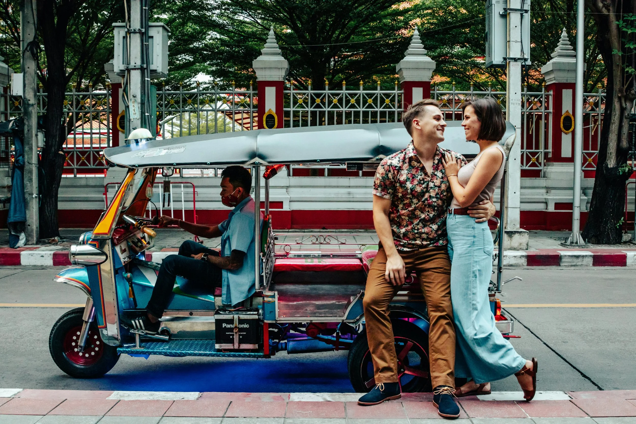 A couple sitting on a tuk-tuk on a city street, smiling at each other while a driver waits behind them.