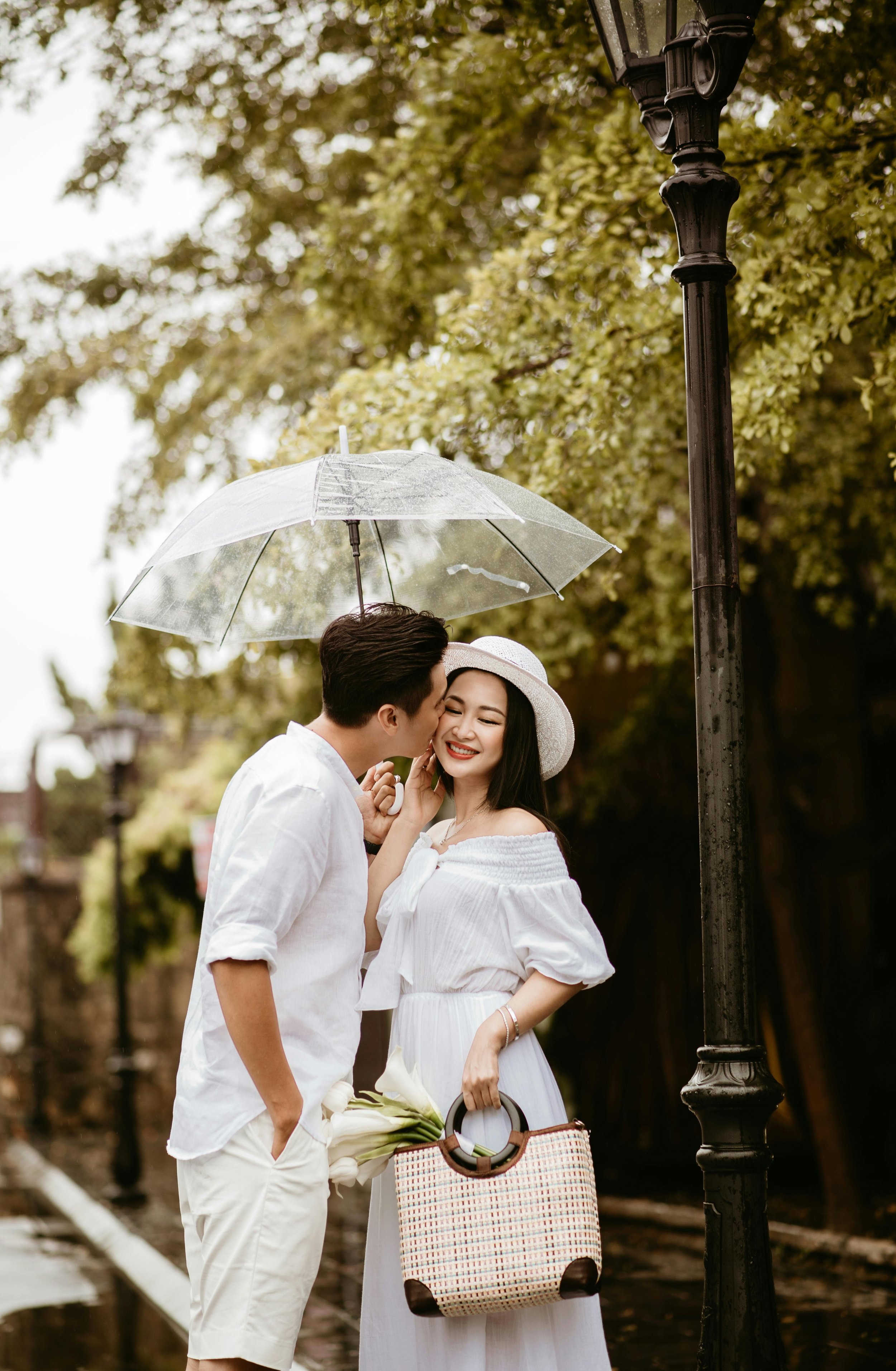 A man and woman under a transparent umbrella sharing a moment, with the man whispering into the woman's ear, both smiling, standing beside a lamppost on a park or outdoor walkway.