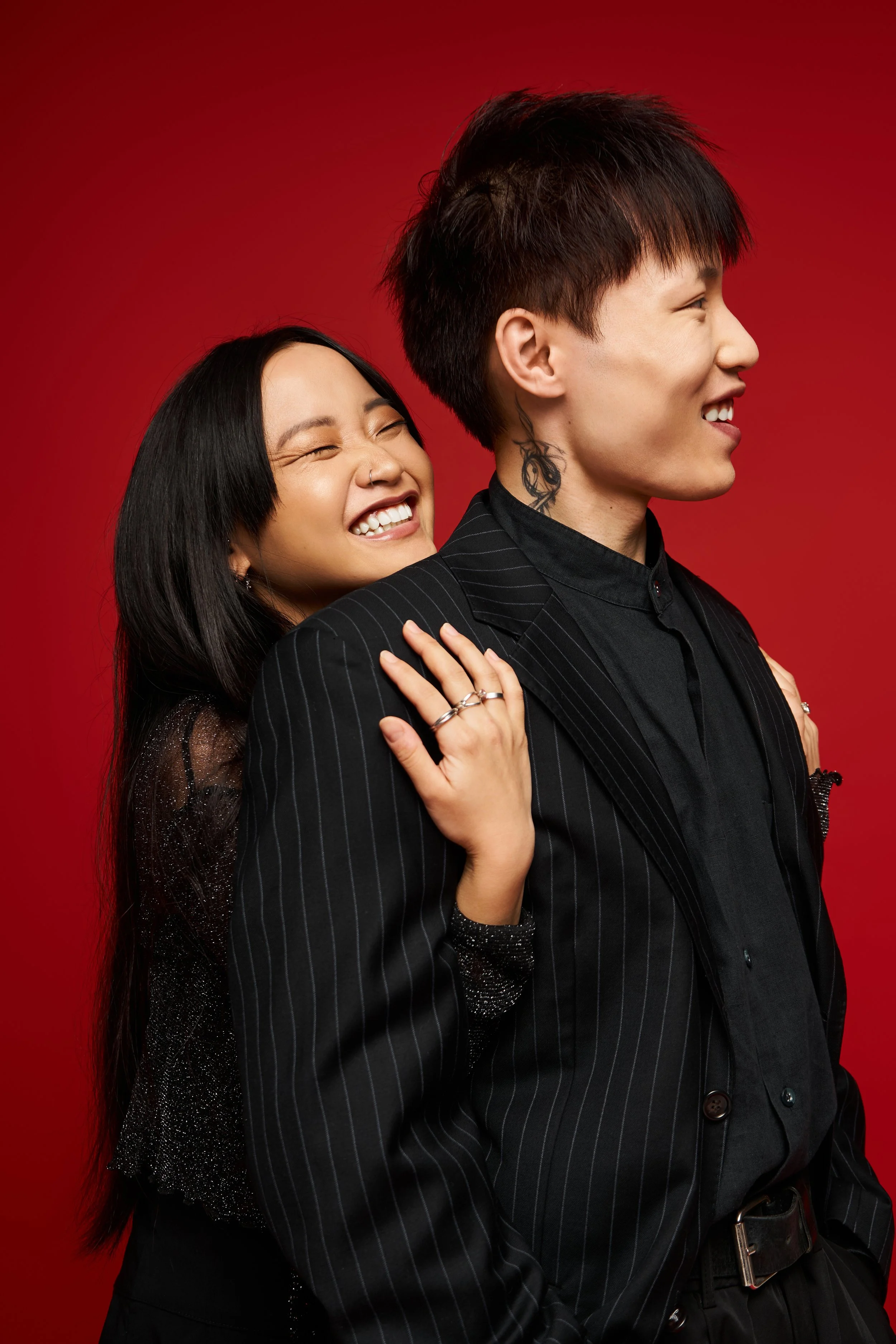 A smiling woman with long black hair hugging a man with short dark hair, who is looking to the side, against a red background.