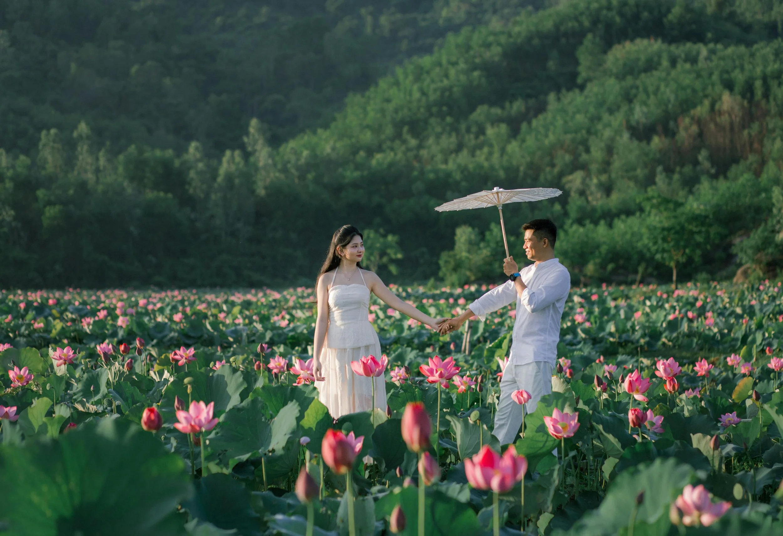 A couple in white clothing standing in a lush field of pink lotus flowers, with a green hillside in the background. The man is holding an umbrella and holding hands with the woman.
