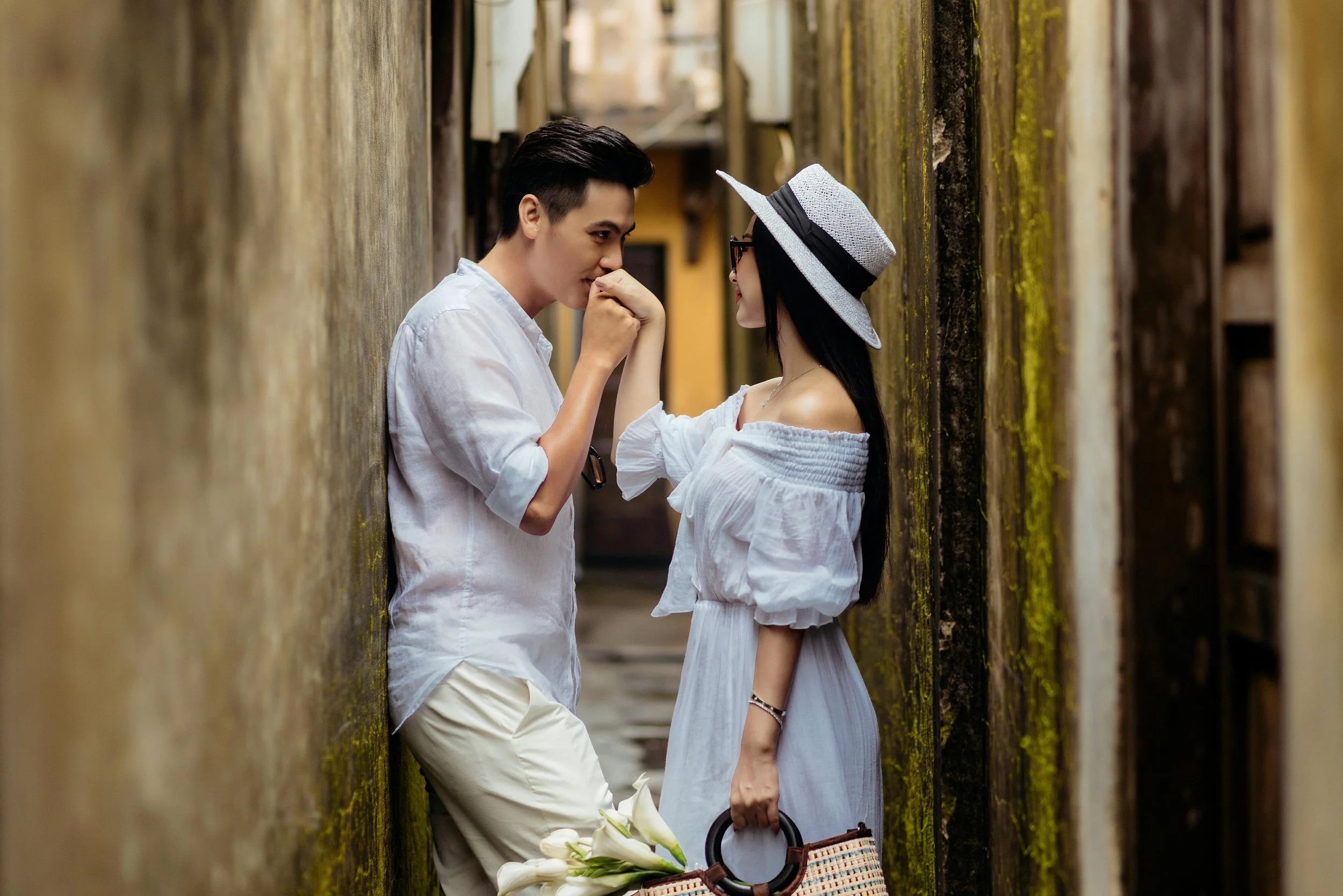 A young man and woman standing close to each other in a narrow alleyway. The man is holding the woman's hand near his mouth, and they are gazing at each other. The woman is wearing a wide-brimmed hat, sunglasses, and a white off-shoulder dress, holdi