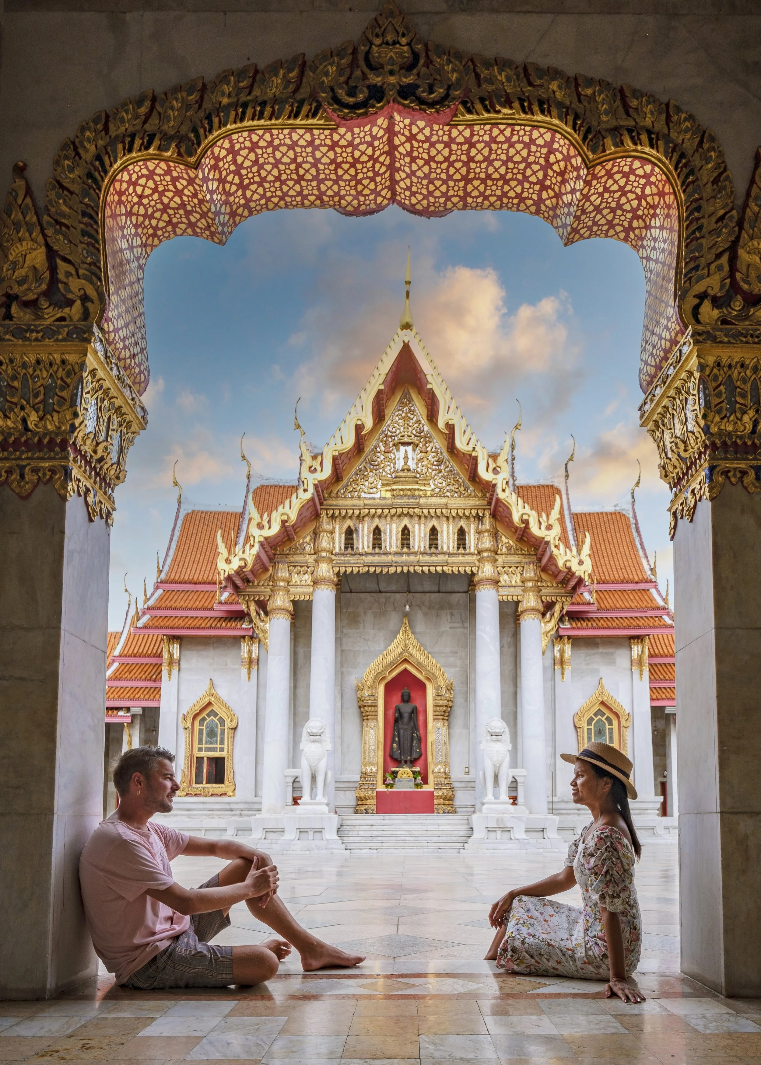 Two people sitting on the floor and talking inside a temple with a golden roof and ornate decorations, with a Buddha statue in the background.