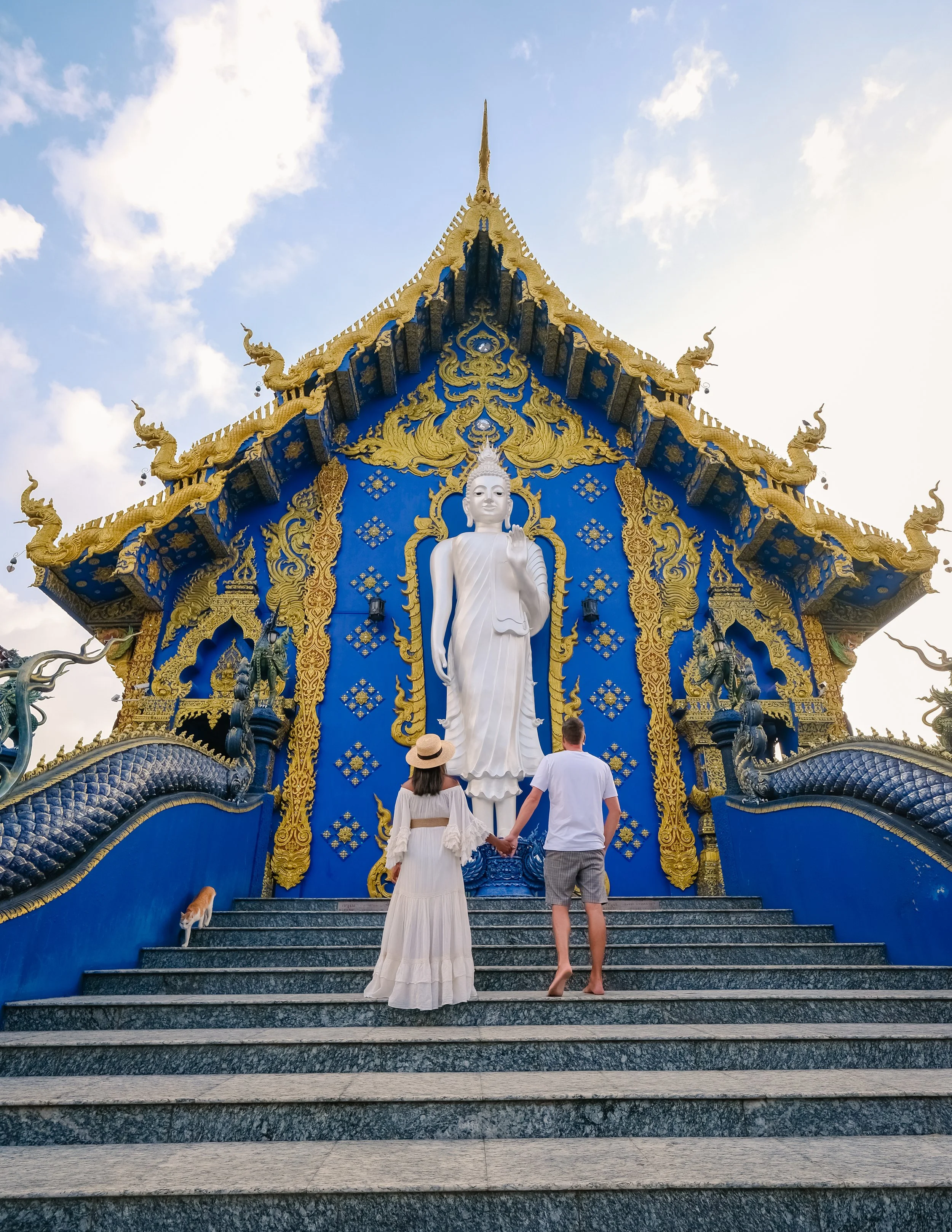 A couple holding hands and walking up stairs towards a large white Buddha statue at a temple with ornate gold and blue architectural detailing.