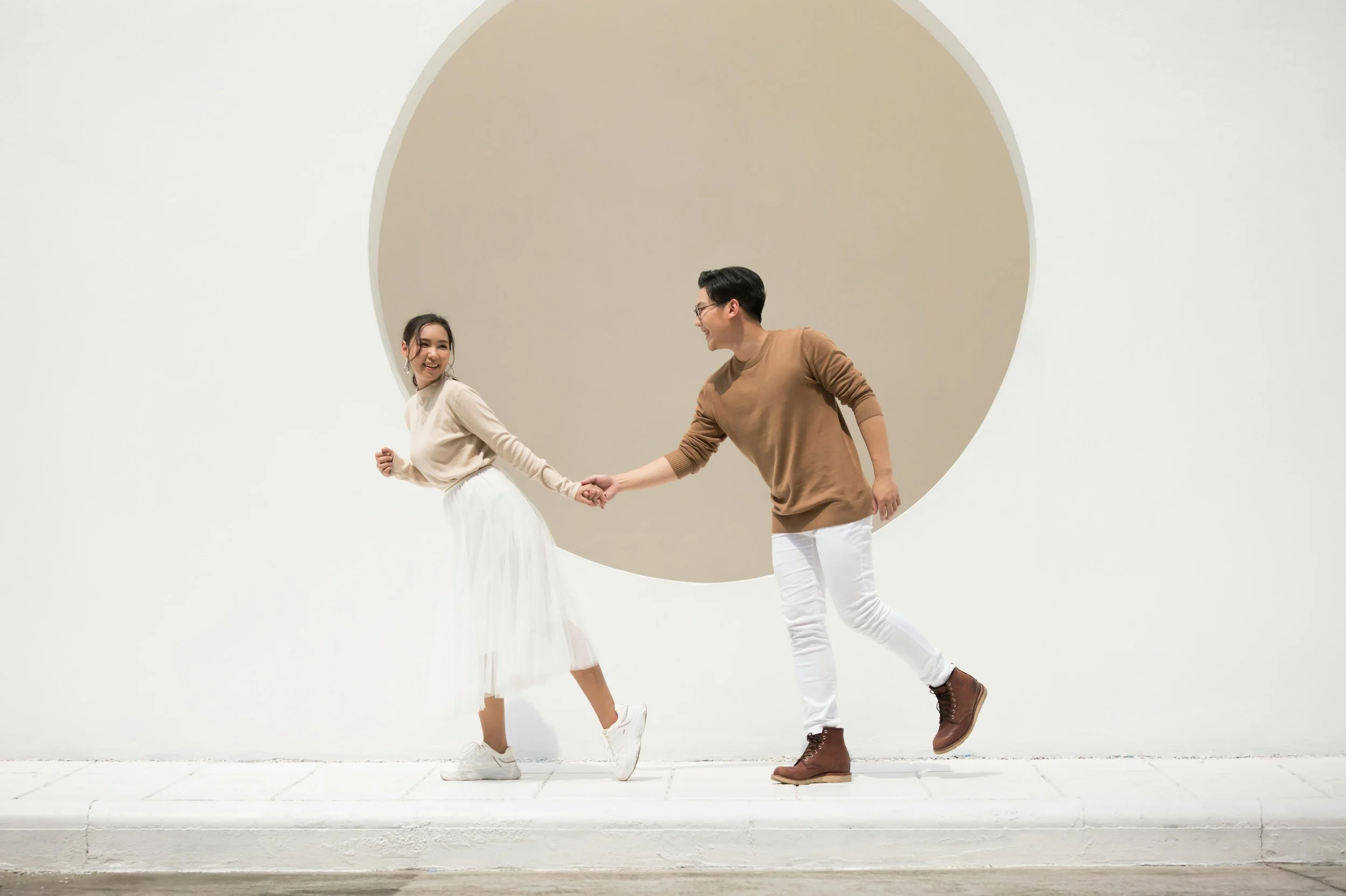 A young man and woman holding hands and smiling on a white platform in front of a large beige circle behind them.
