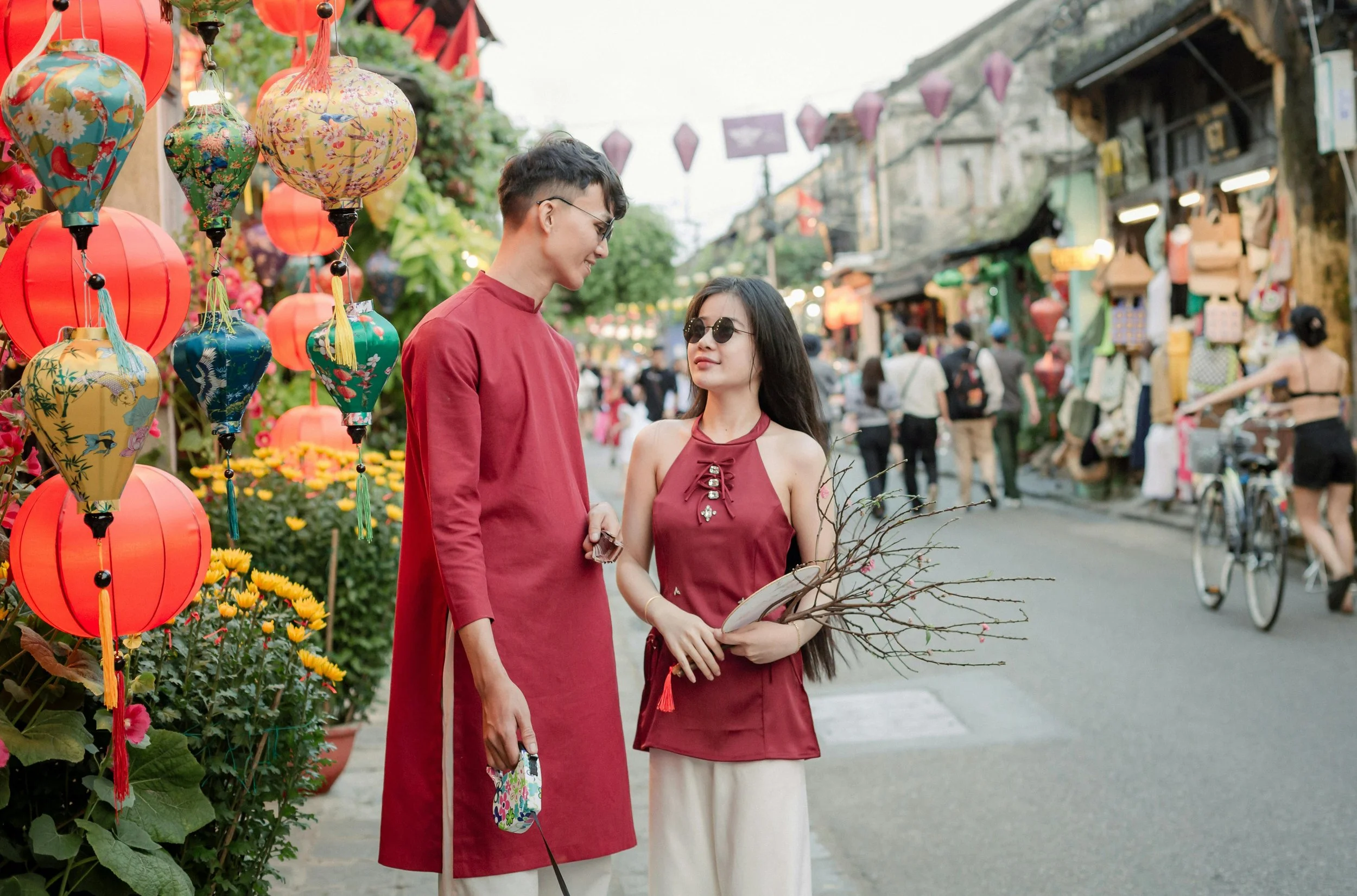 A young man and woman in traditional Vietnamese clothing standing on a busy street decorated with colorful lanterns and flowers during daytime.