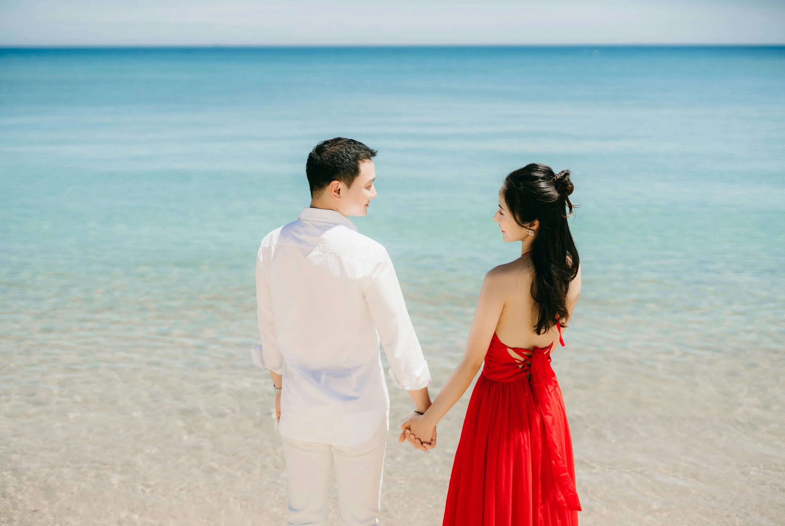 A couple holding hands on a beach, facing each other and smiling, with the ocean behind them and a clear sky above.