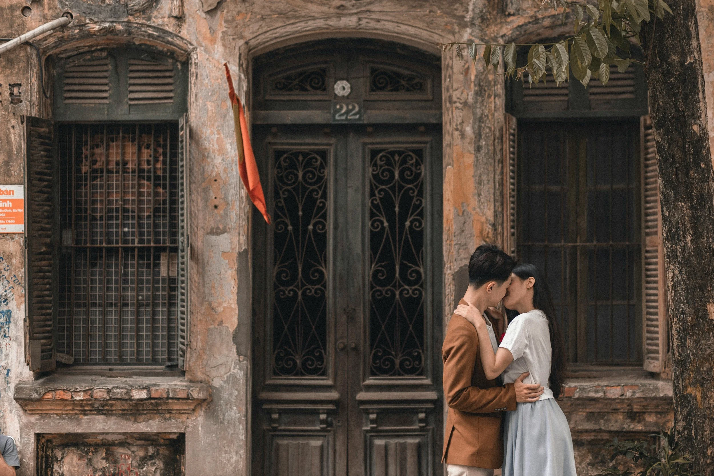 A young couple kissing in front of an old, weathered building with graffiti and wooden shutters.