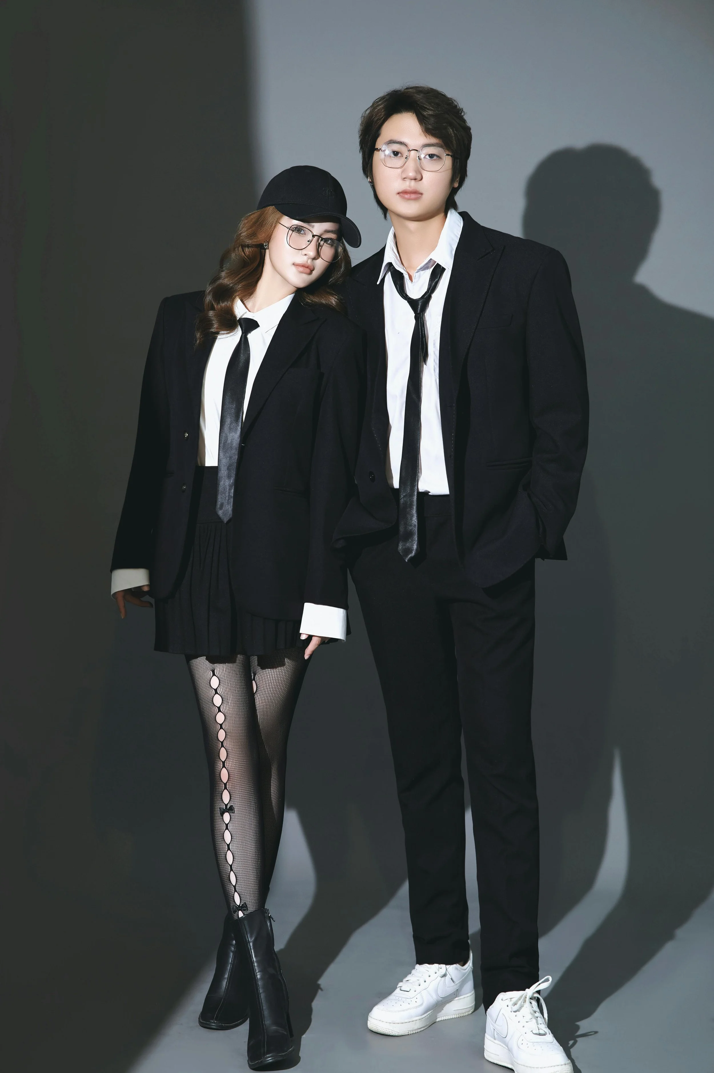 A man and woman in black suits and ties standing against a dark gray background, with the woman wearing fishnet tights and boots, and the man wearing white sneakers.
