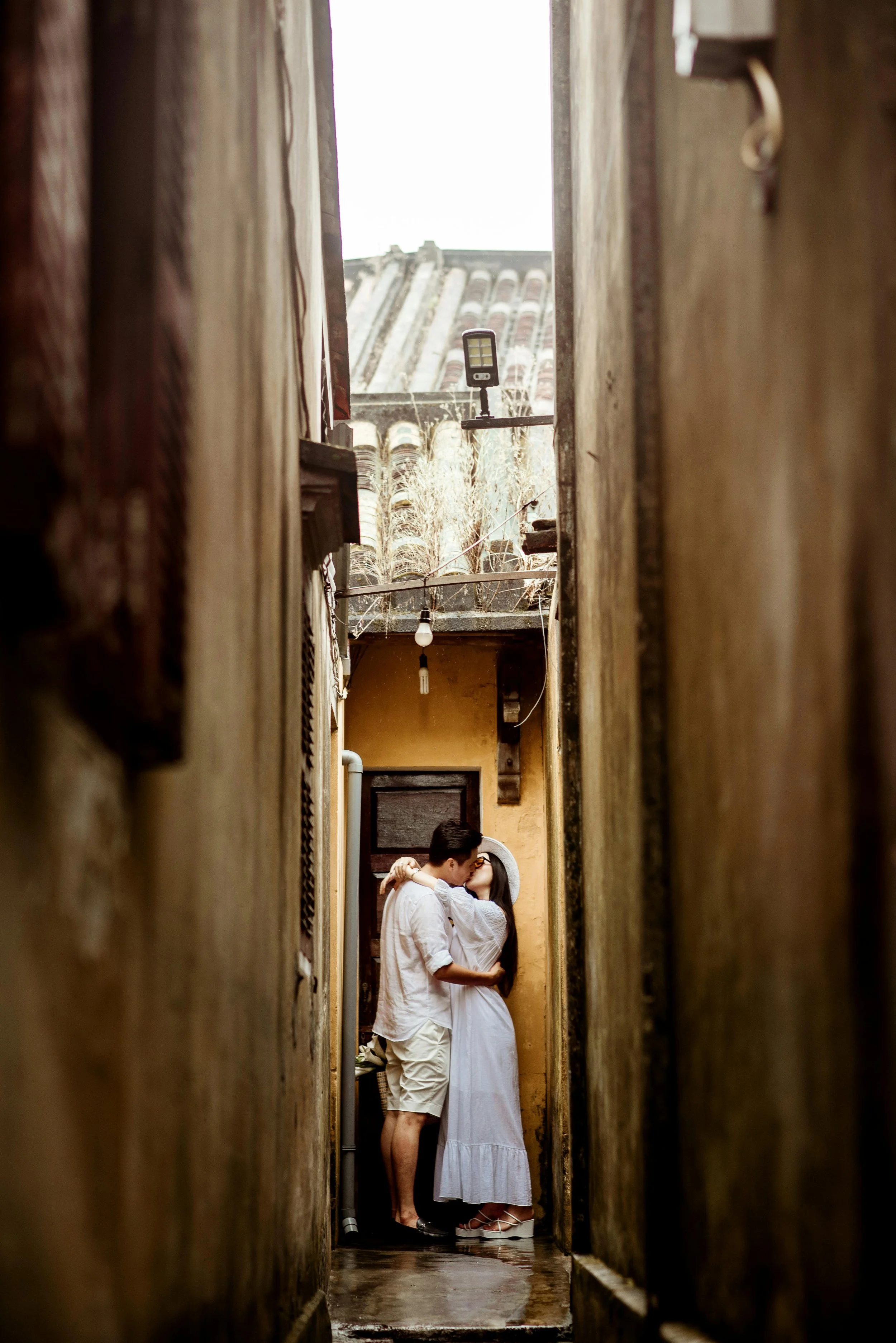 A couple standing in a narrow alley, kissing, with the man in a white shirt and shorts, and the woman in a long white dress and wide-brimmed hat, surrounded by old building walls.