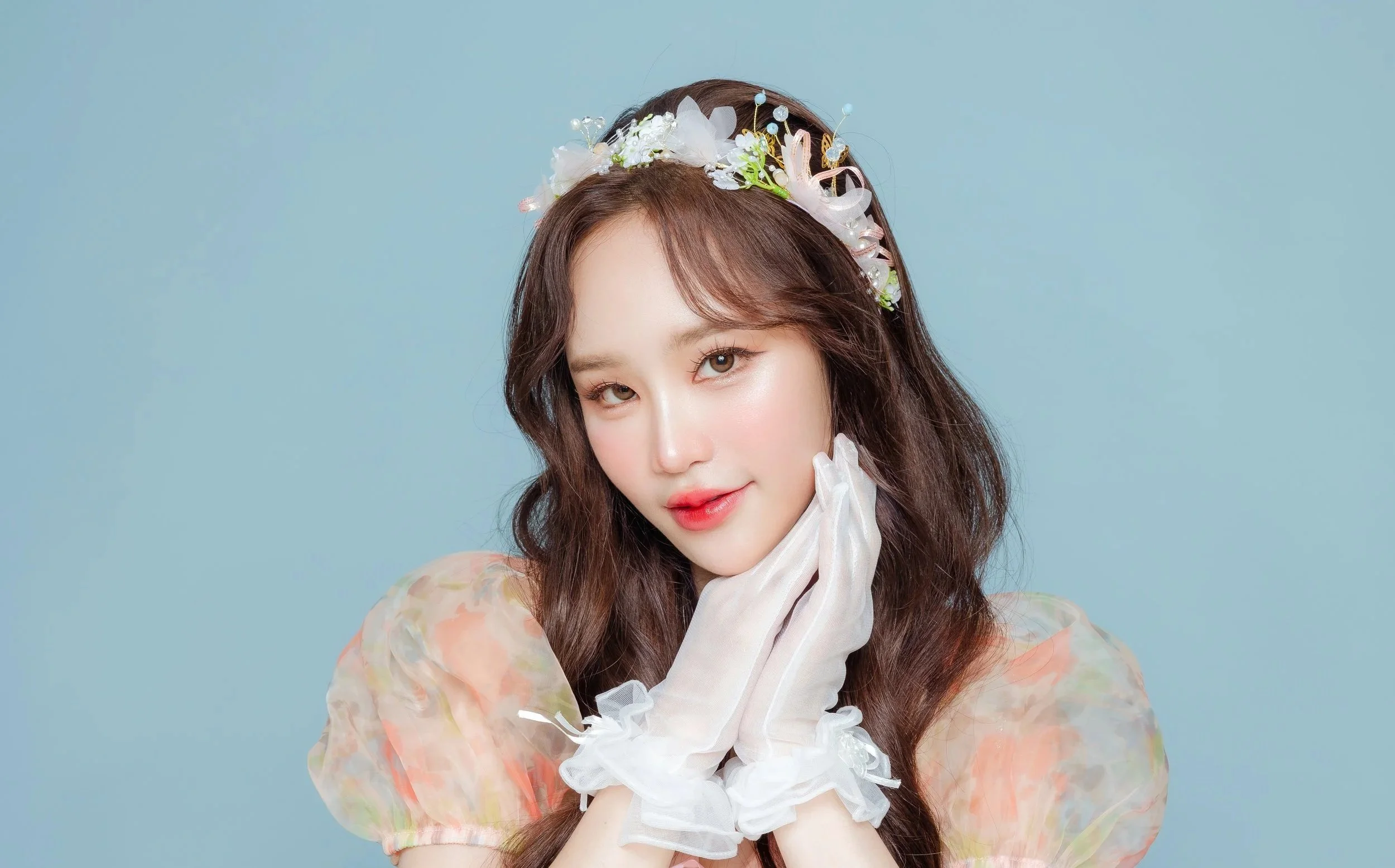 A young woman with long brown hair wearing a floral headband and sheer pastel-colored puffed sleeves, posing with her hands gently touching her face against a light blue background.