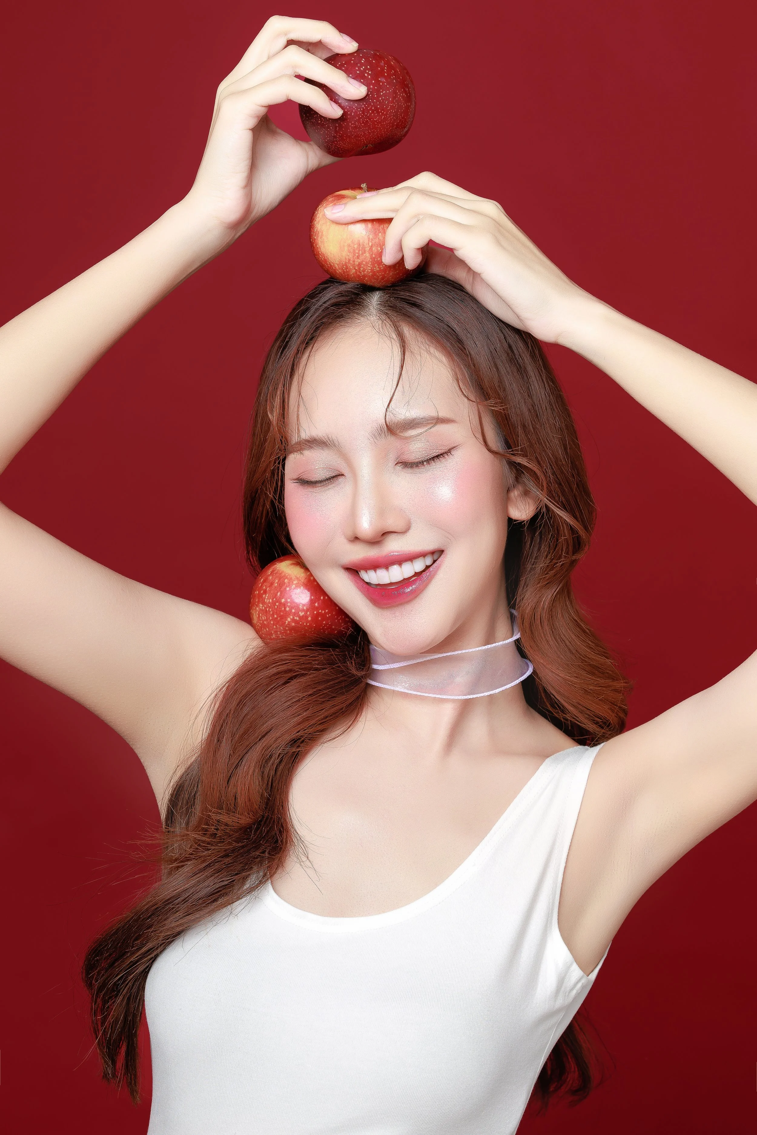 A young woman with long brown hair and a white sleeveless top, smiling with eyes closed, balancing three red apples, one on her shoulder, one on her head, and one near her face, against a red background.