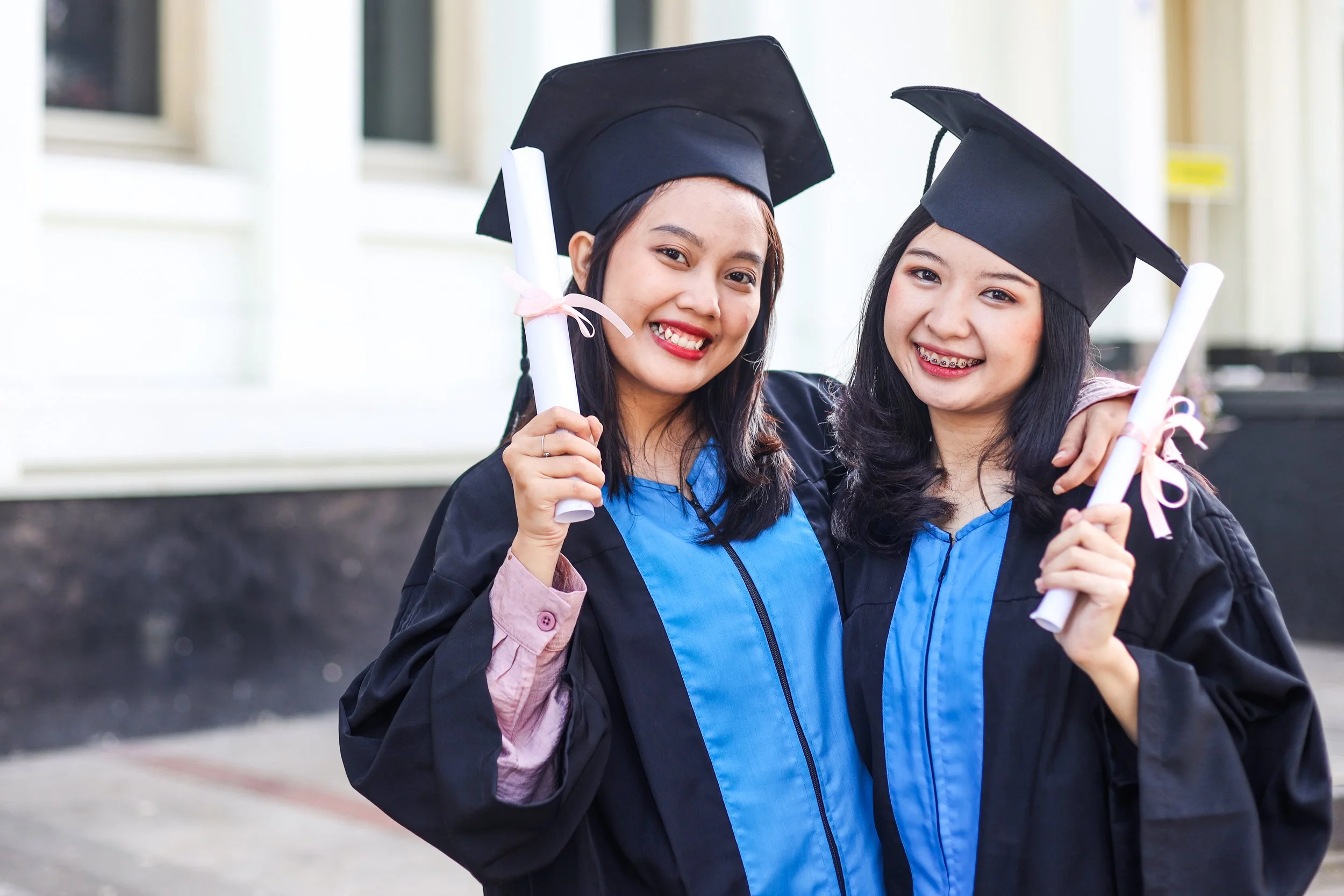Two young women in graduation gowns and caps smiling and holding diplomas.
