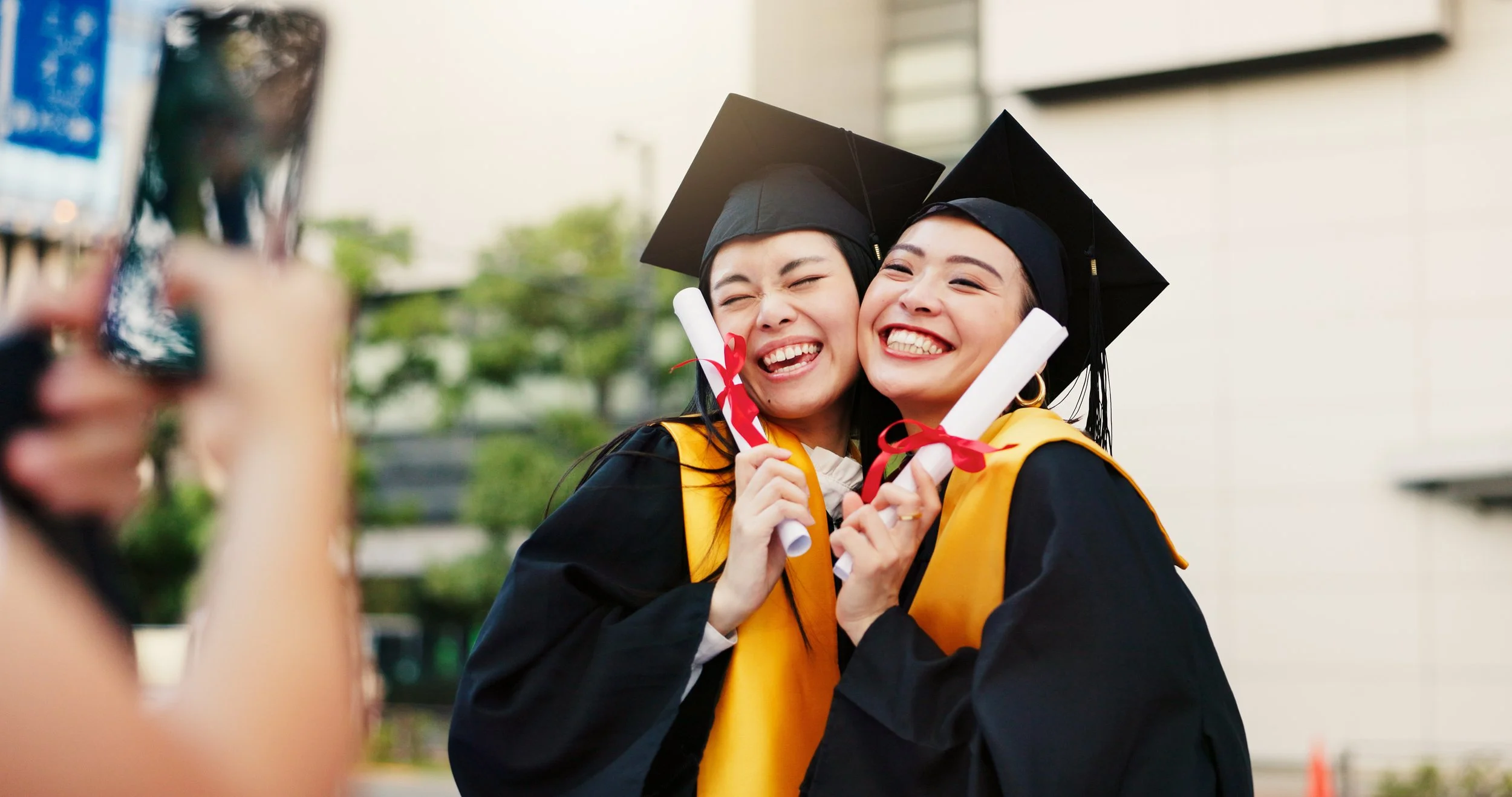 Two women in graduation caps and gowns smiling and hugging, holding diplomas, during graduation ceremony.
