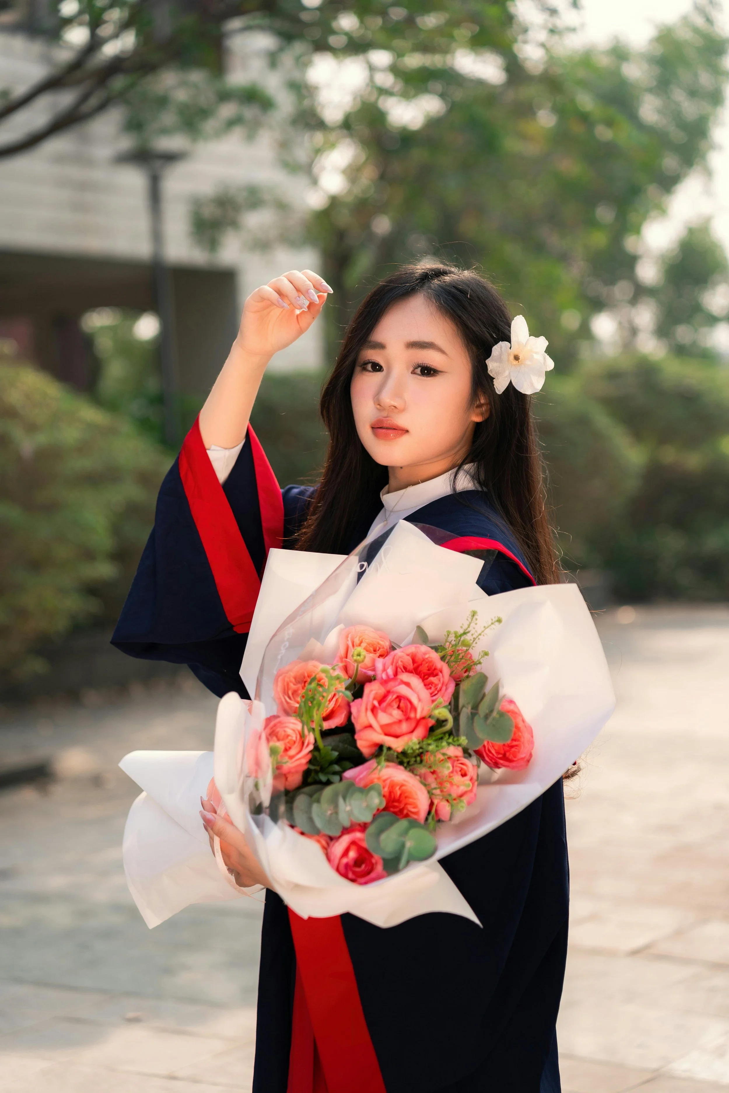 Young woman in graduation gown holding a bouquet of pink flowers, standing outdoors with trees in the background.