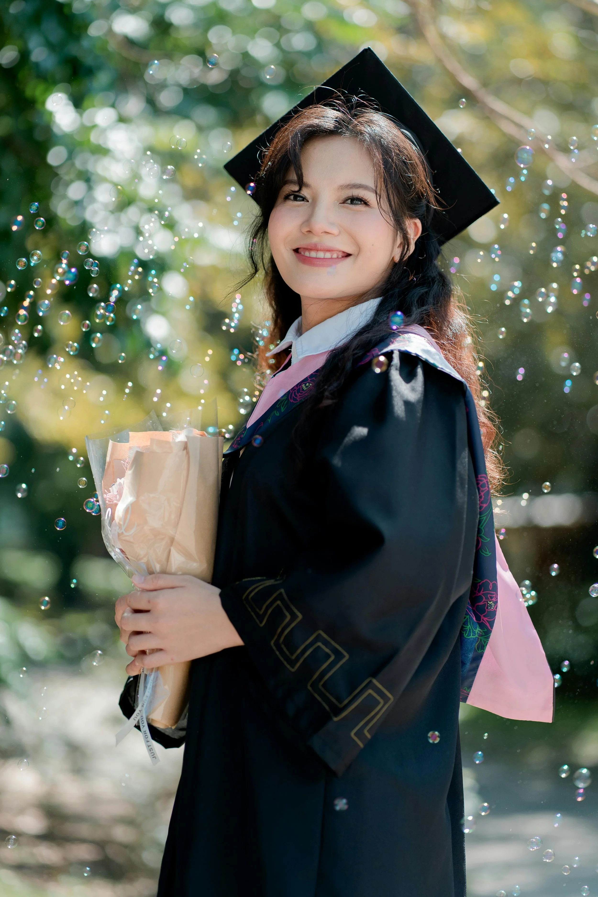 A young woman in a graduation cap and gown holding a bouquet of flowers outdoors with blurred green foliage and bubbles around her.