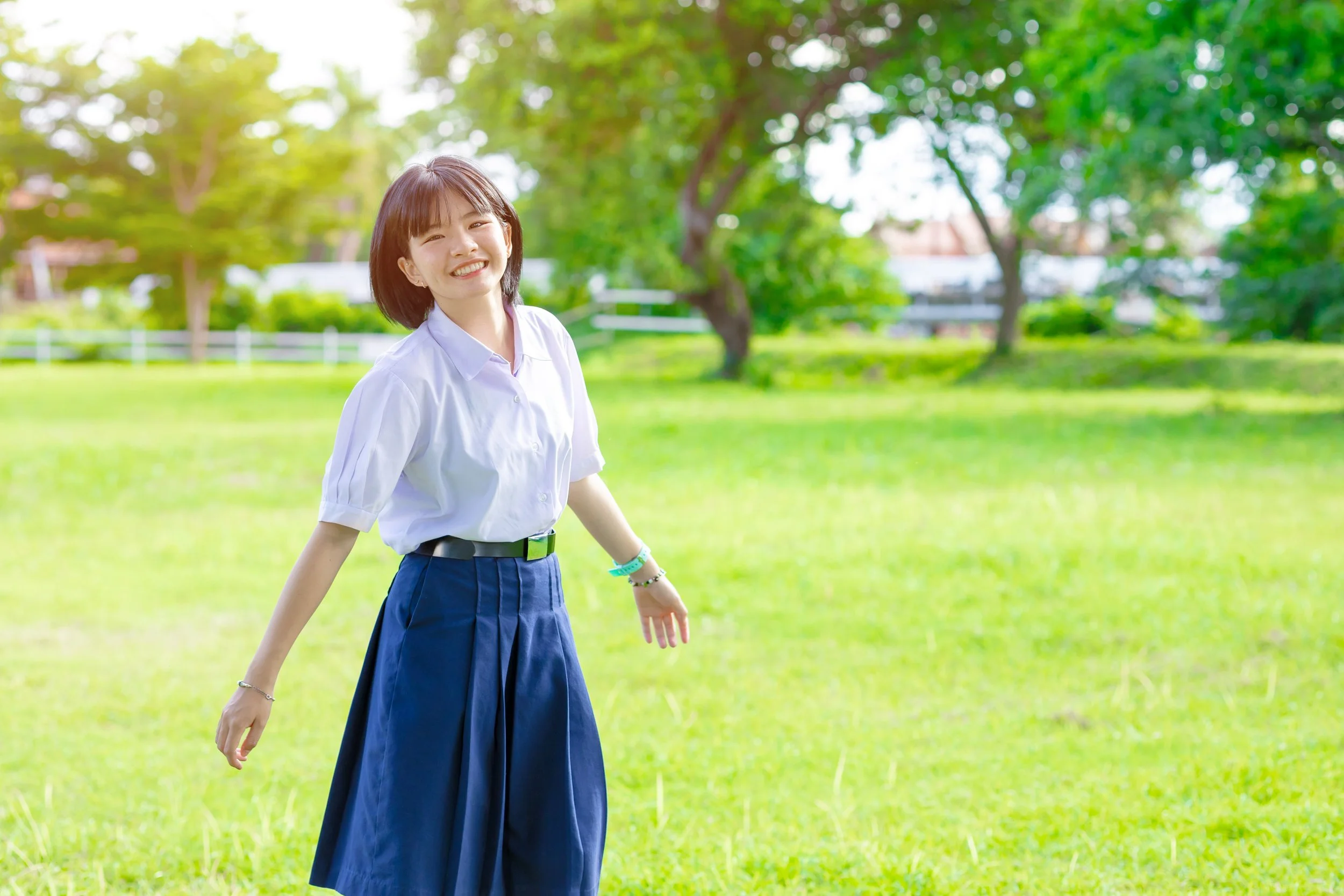 A young woman with short dark hair and a broad smile, wearing a white shirt and a blue skirt, is standing in a lush green park on a sunny day, with trees and grass in the background.