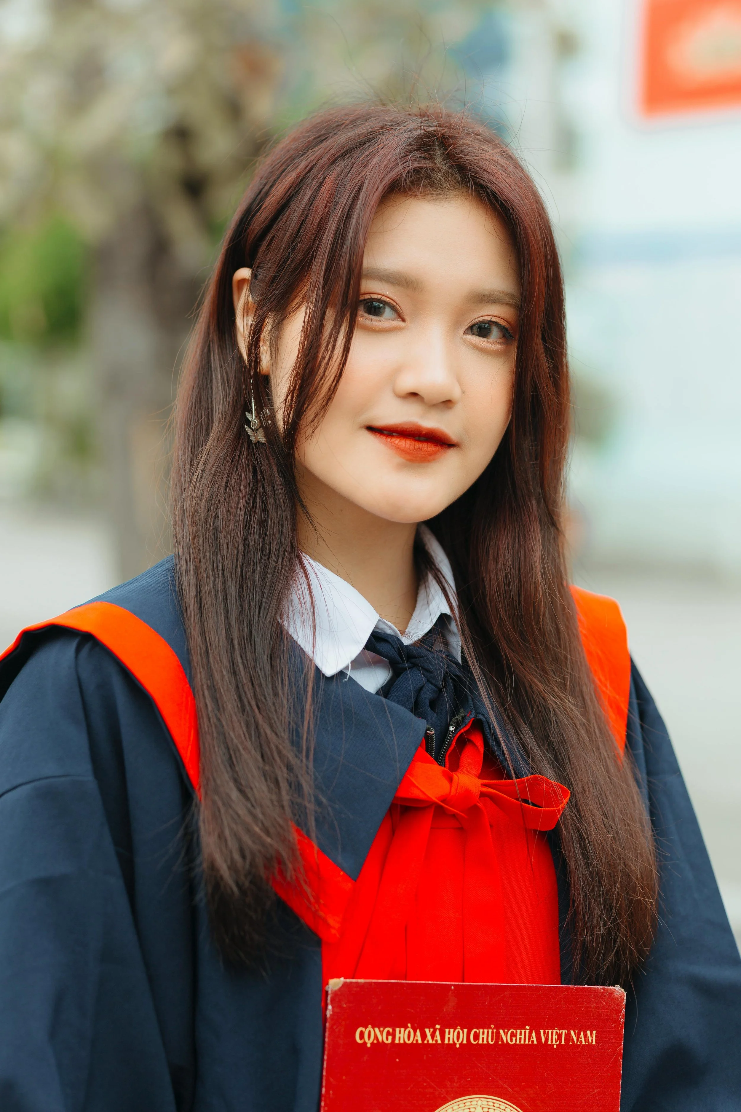 Young woman in graduation gown holding a red diploma with Vietnamese text, outdoors, smiling slightly.