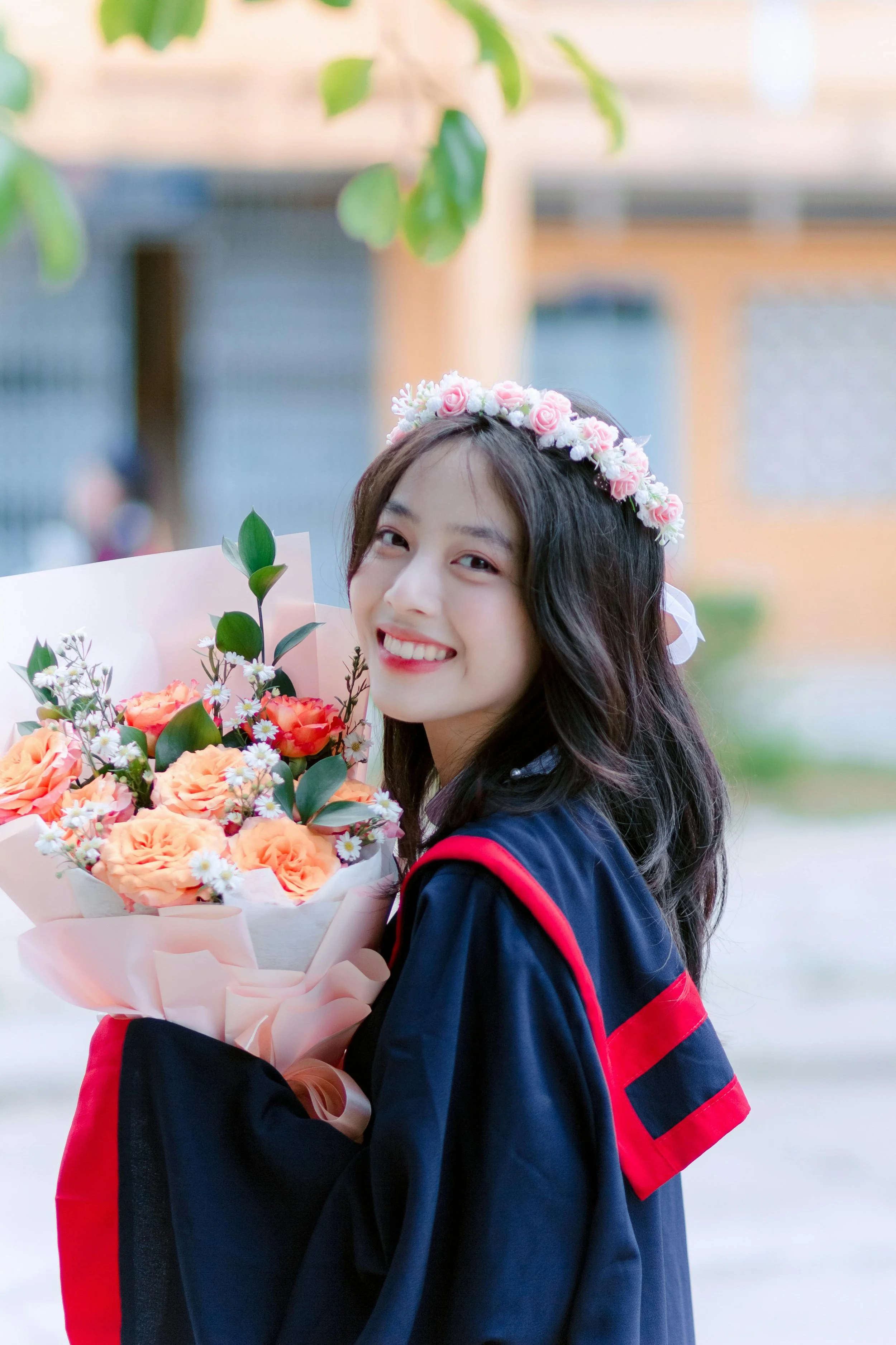 Young woman in a graduation gown holding a bouquet of peach and white flowers, wearing a pink floral headband, smiling outdoors.
