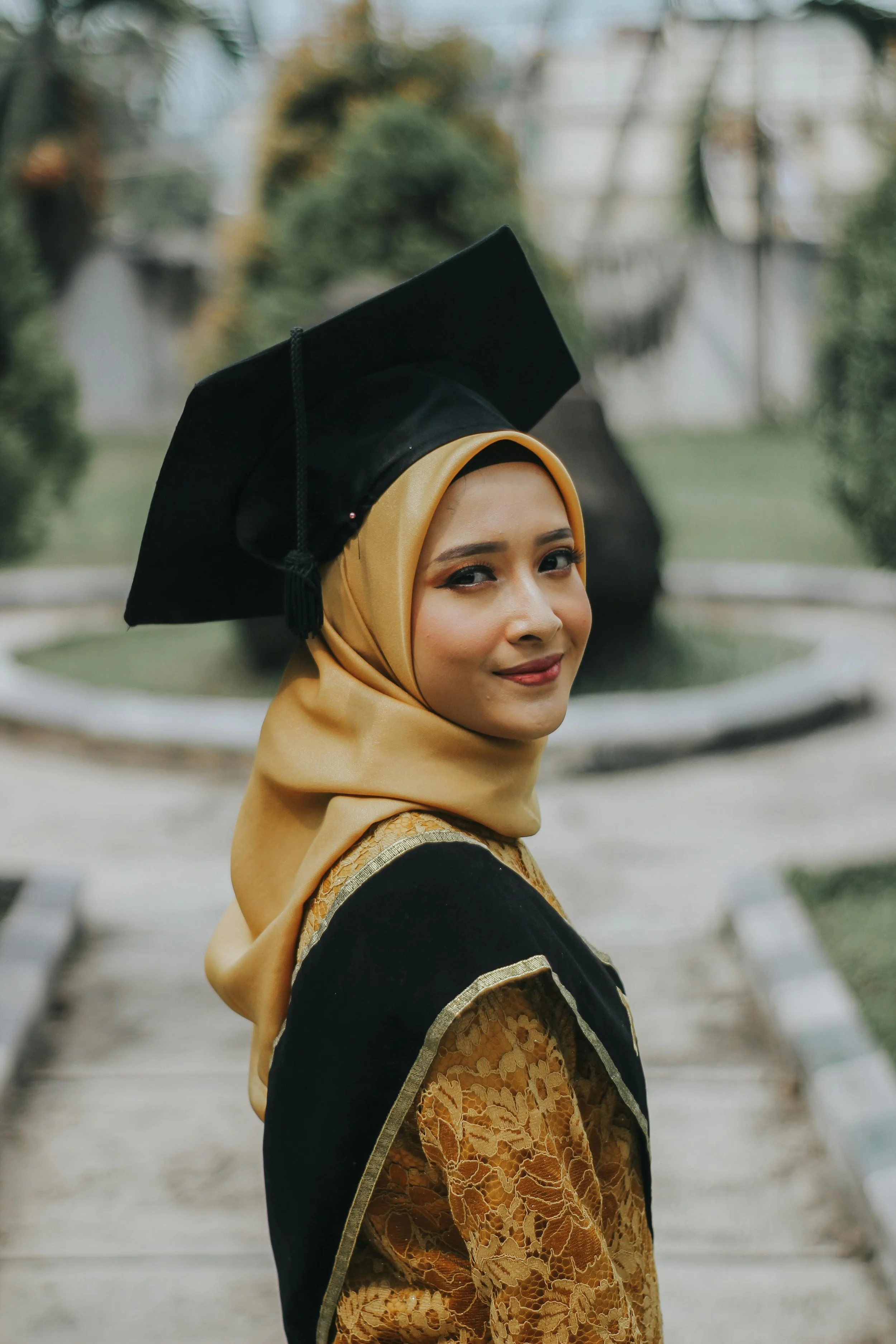 A young woman wearing a graduation gown and cap, with a beige hijab, standing outdoors near a fountain, smiling at the camera.