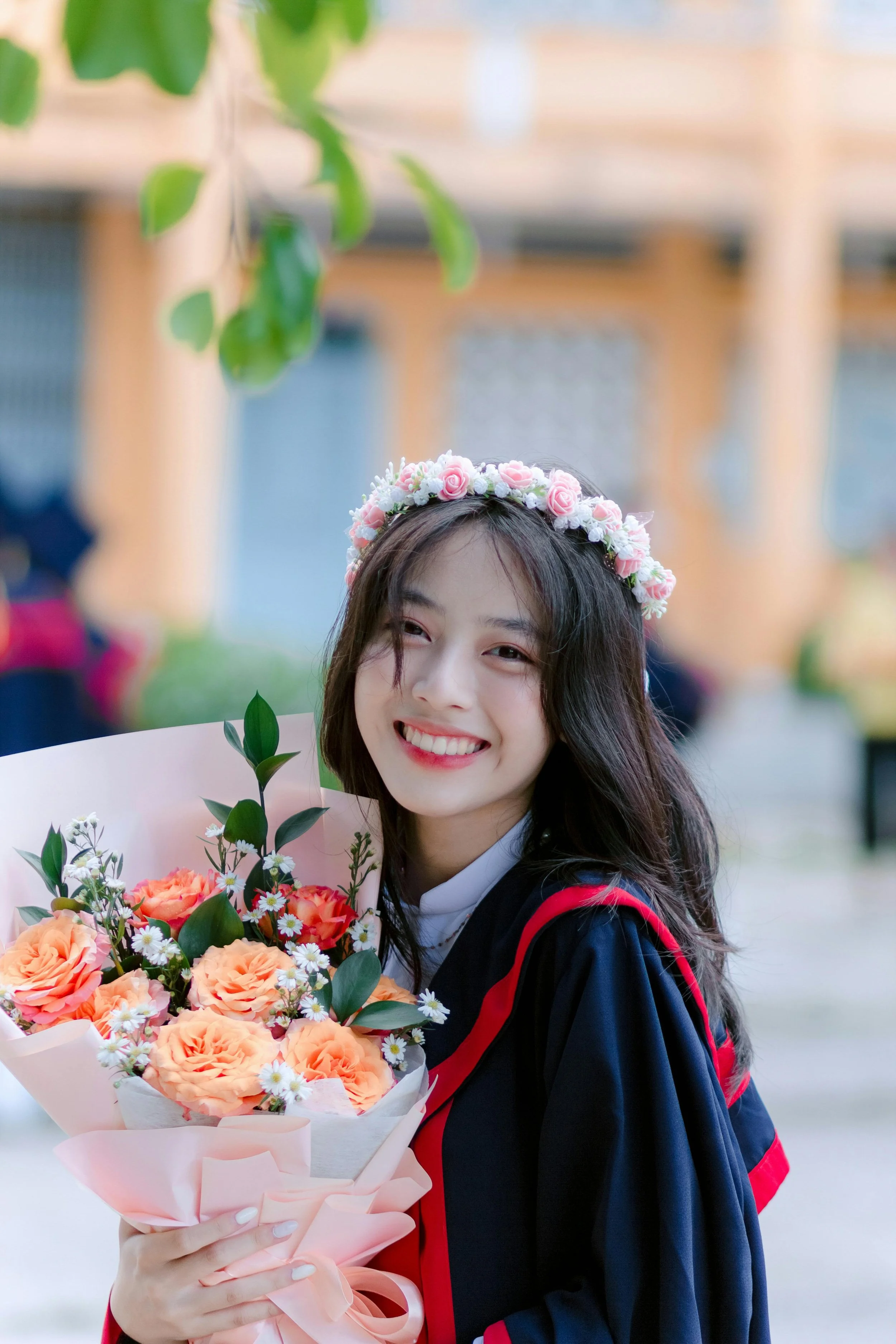A young woman in a graduation gown holding a bouquet of flowers and wearing a flower crown, smiling at a graduation ceremony.