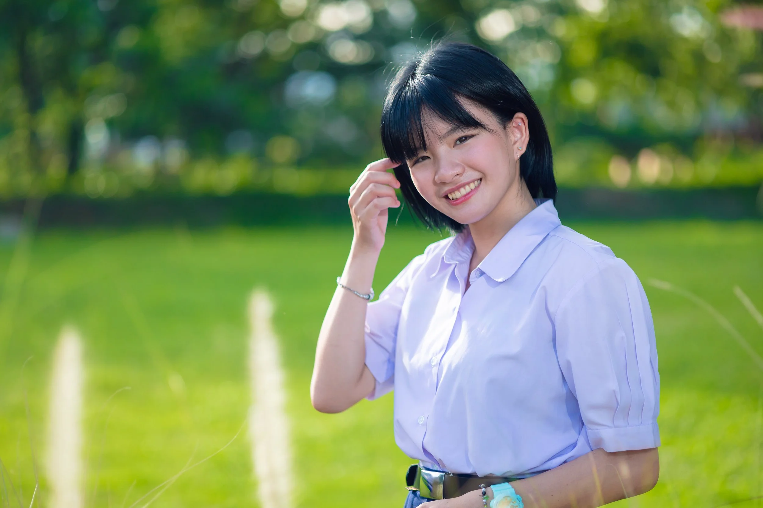 A young woman with short black hair, wearing a white short-sleeved button-up shirt, smiling outdoors in a lush green park on a sunny day.