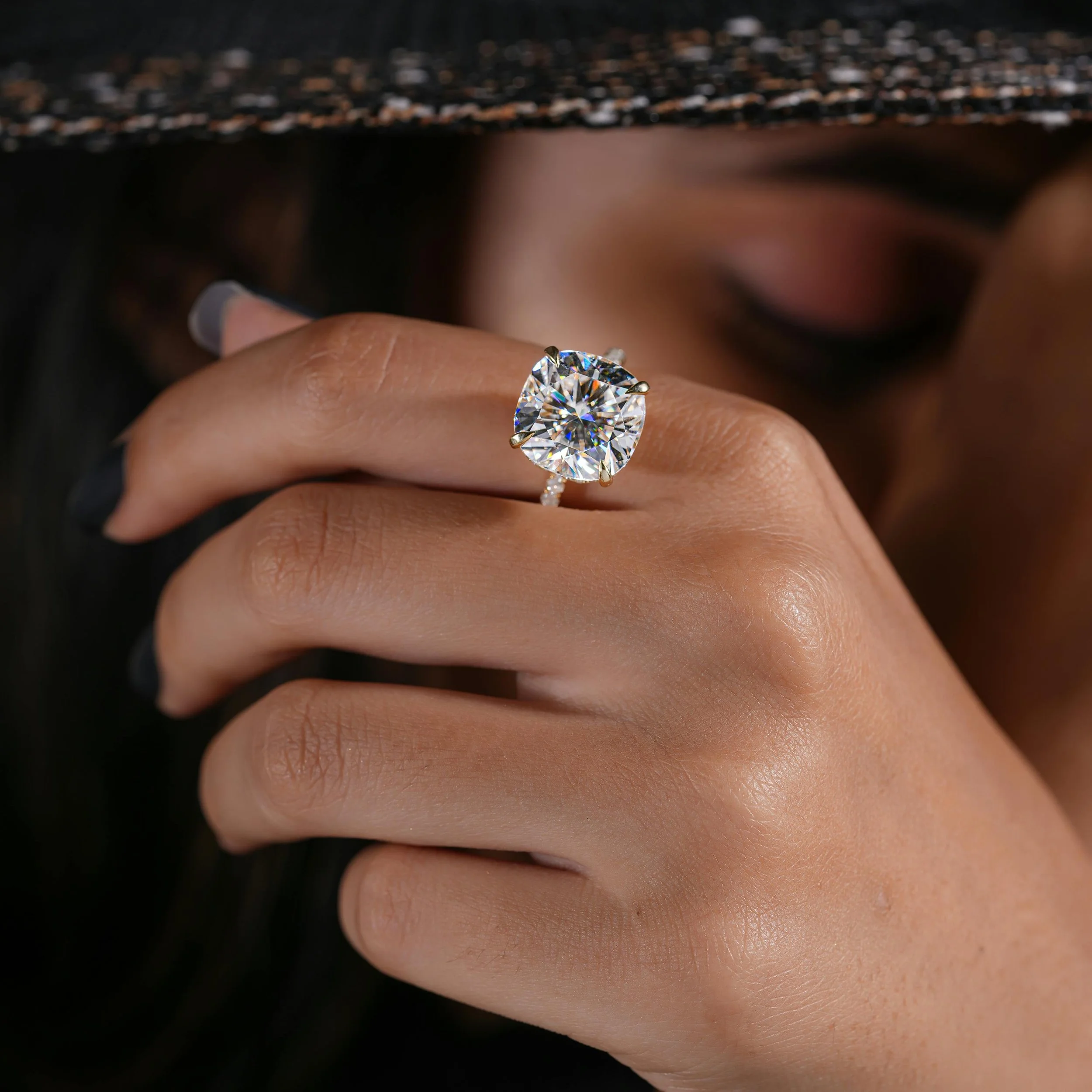 Close-up of a woman wearing a large diamond engagement ring with a cushion-cut stone on her finger, with her face partially visible in the background.