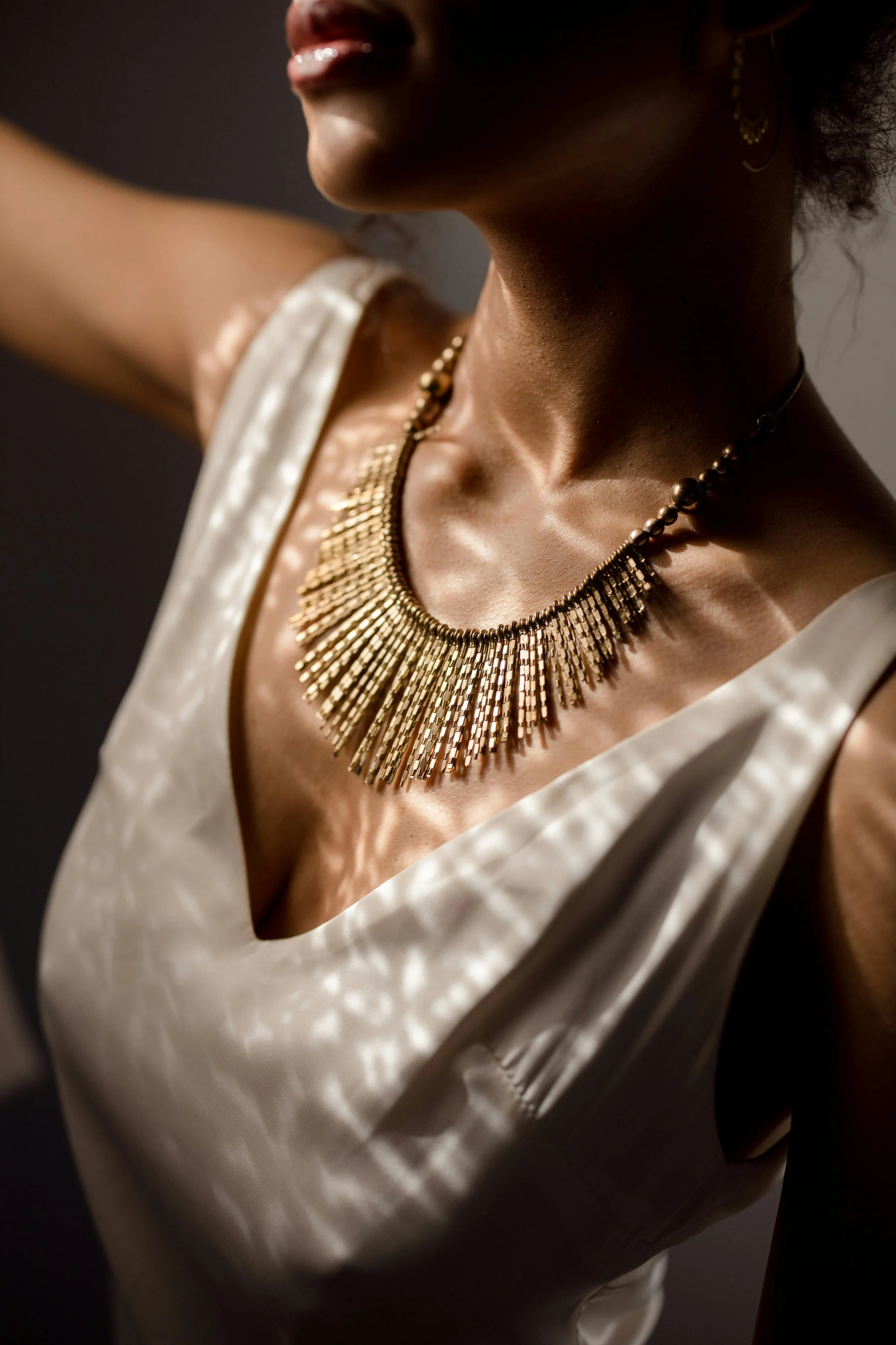 Close-up of a woman wearing a white satin dress and a large ornate gold necklace. The focus is on her neck, collarbone, and part of her face, with shadows cast by her jewelry and clothing.