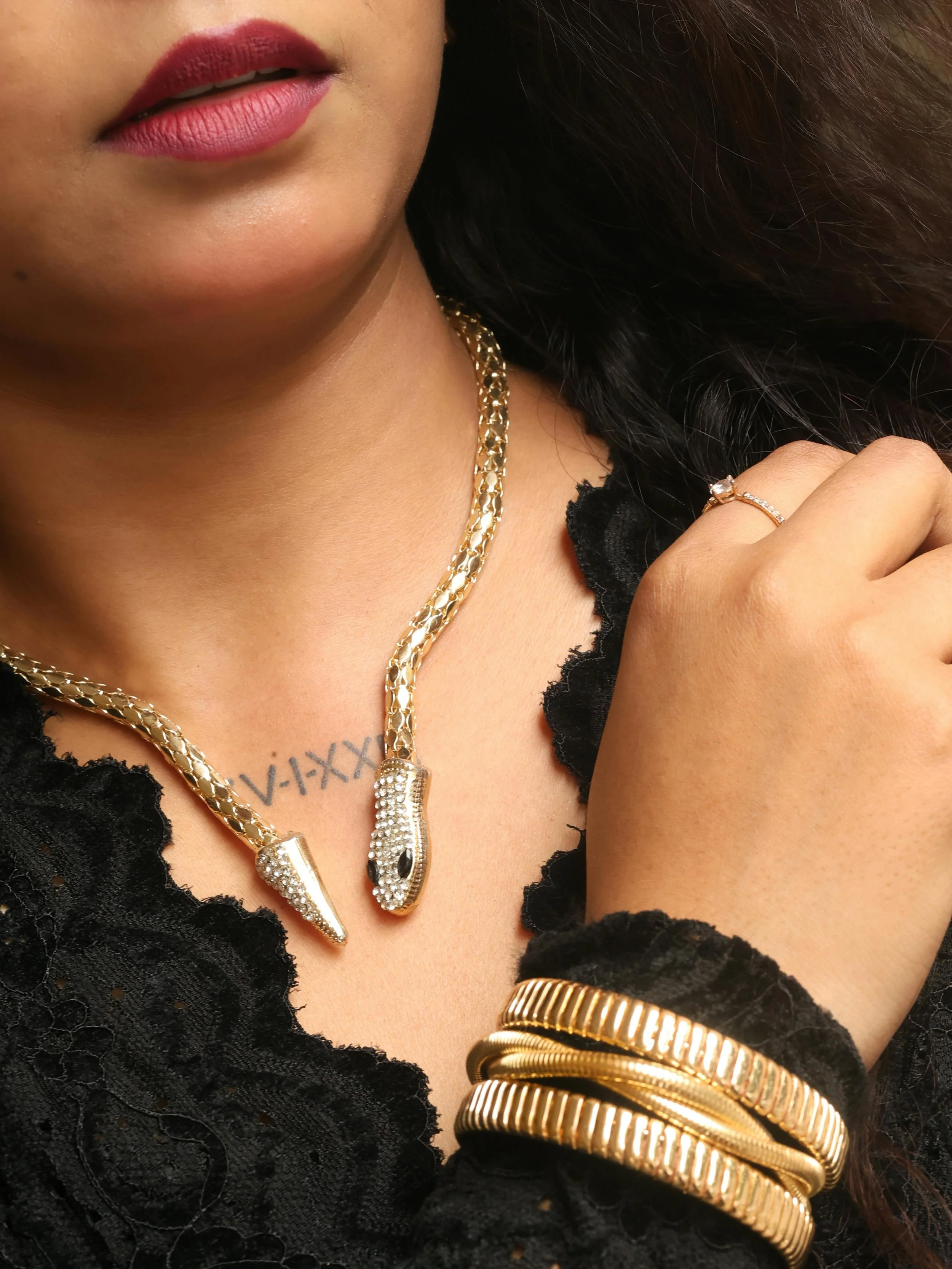Close-up of a woman's neck and hand showing gold jewelry including a snake-shaped necklace with diamond accents, a ring with a central diamond, and multiple gold bangles; part of her lips and black lace clothing are also visible.