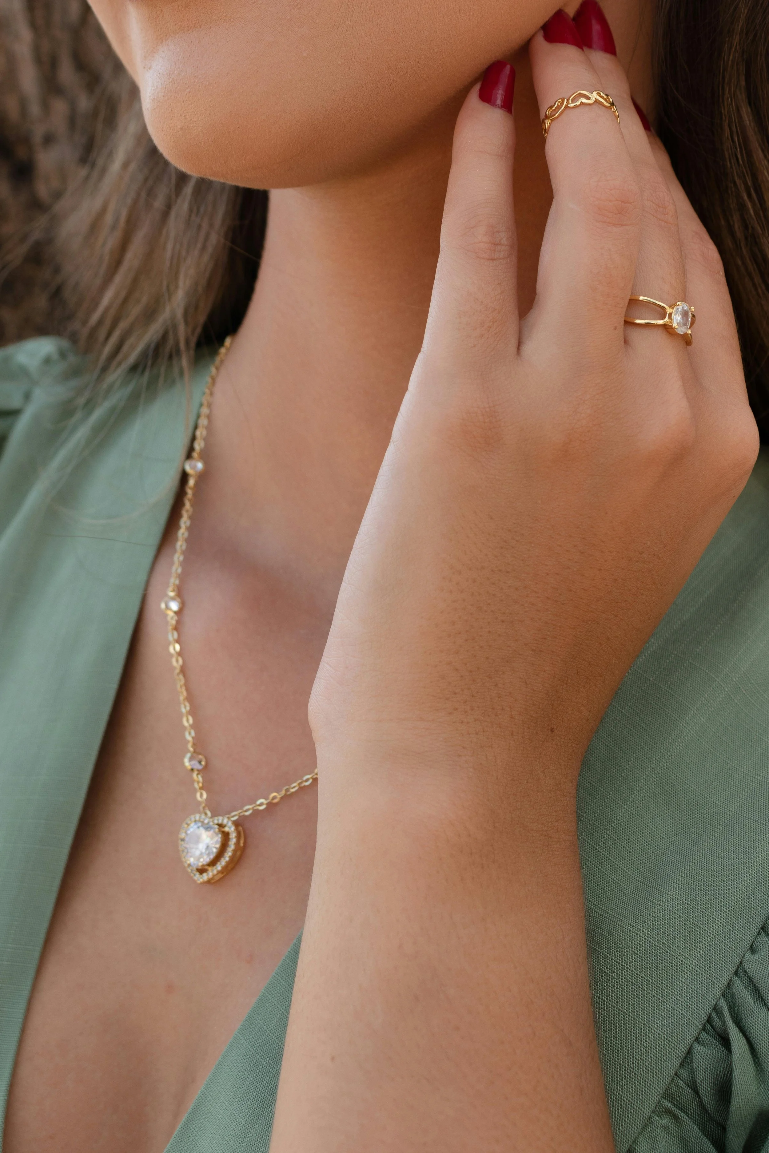 Close-up of a woman's hand with gold jewelry, including a ring and a ring, touching her face. She is wearing a necklace with a large heart-shaped pendant and a green top.