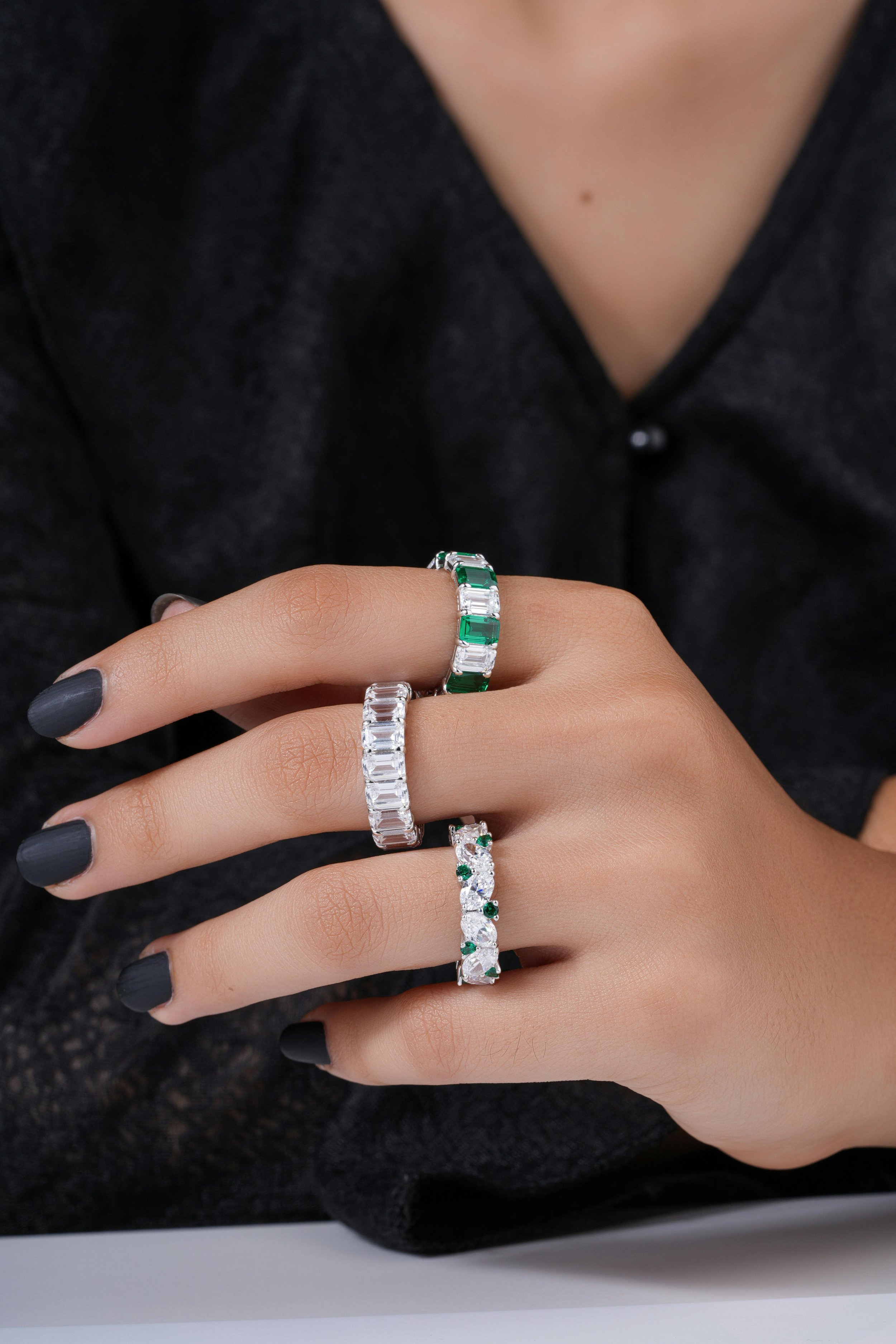 Close-up of a woman's hand with three silver rings featuring emerald and diamond accents, against a black shirt background.