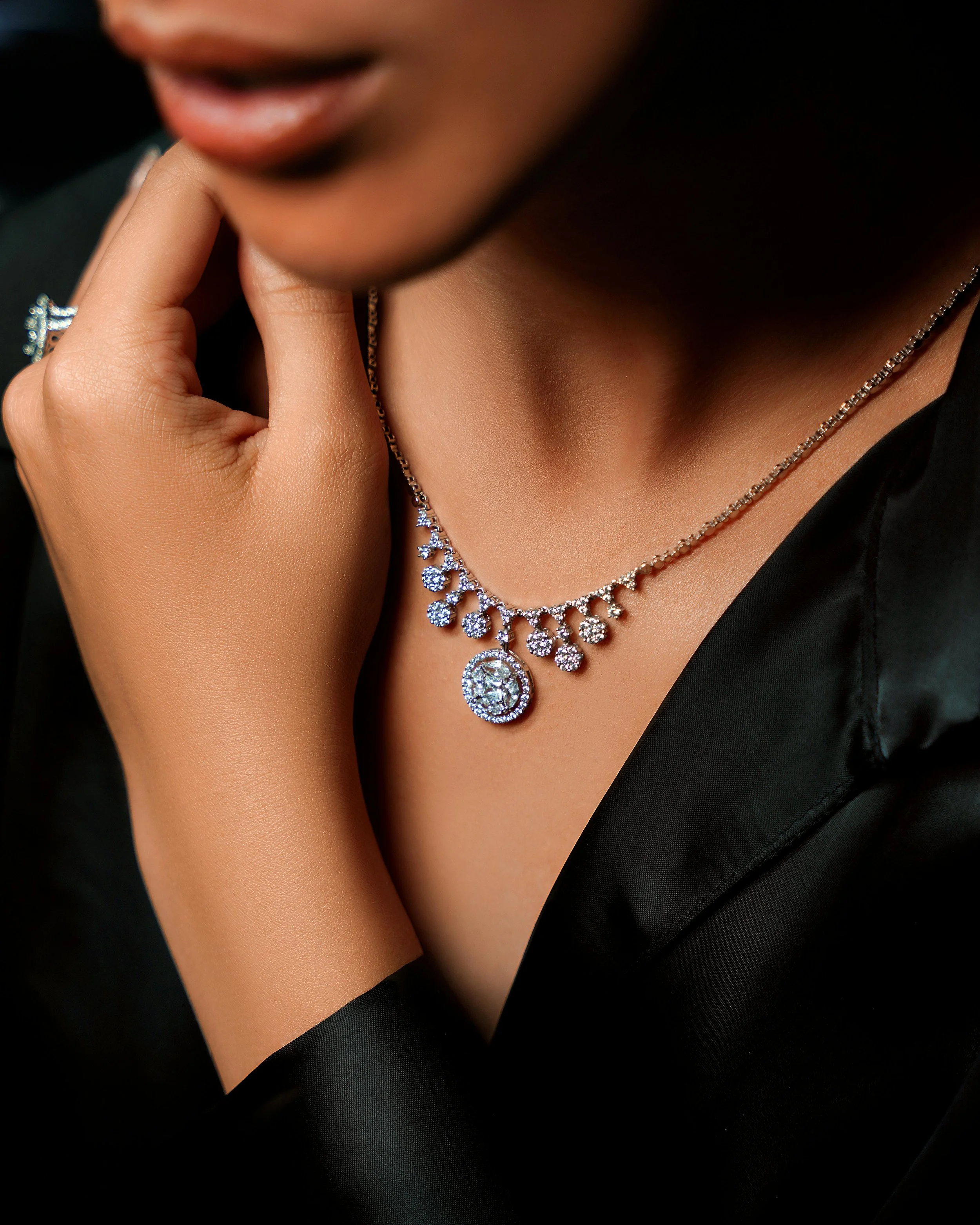 A woman wearing an elegant diamond necklace with a circular pendant, touching her chin with her hand, dressed in a black satin garment.
